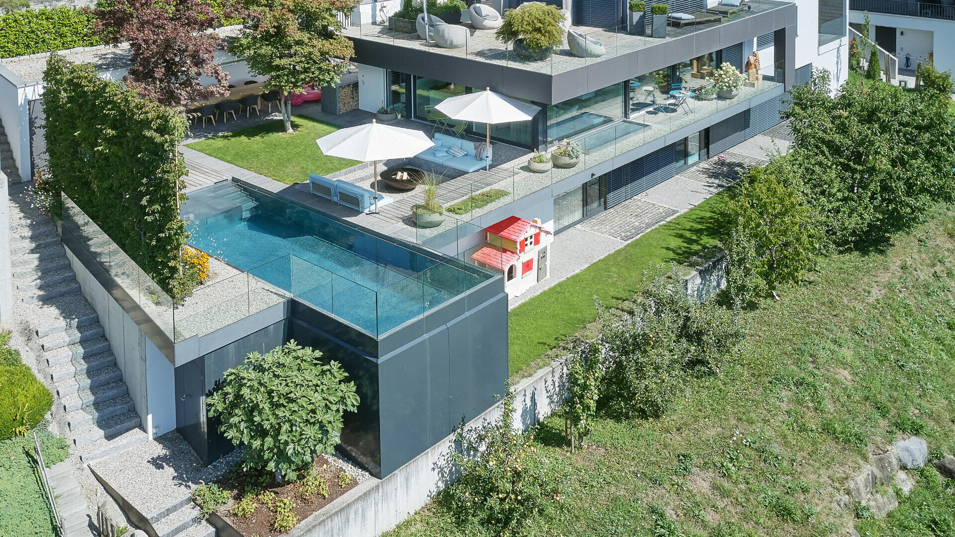 Modernes mehrstöckiges Haus mit einem Dachgarten, großen Glasfenstern, einem Außenpool mit Glaswänden, Liegestühlen, Sonnenschirmen und einem kleinen rot-weißen Spielhaus auf einer grünen Wiese.