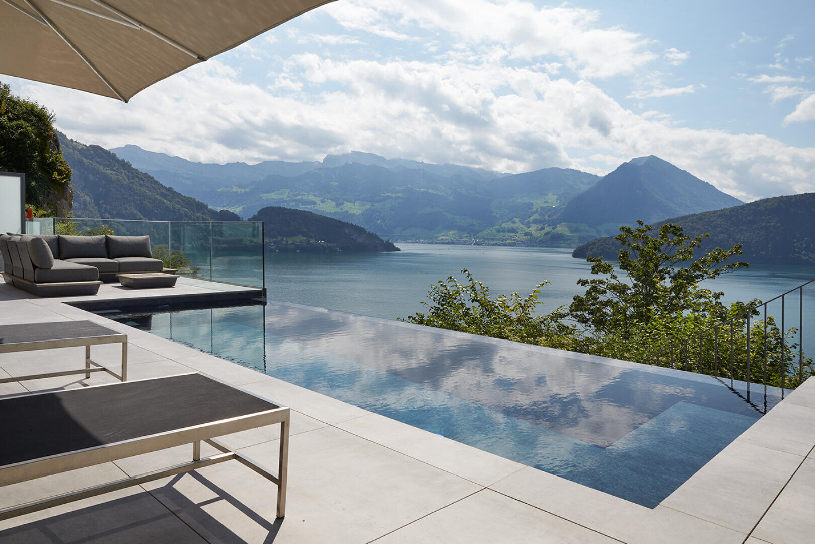 Infinity-Pool mit Blick auf einen ruhigen See und die Berge, mit modernen Liegestühlen und einem Sofa auf einer sonnenbeschienenen Terrasse. Ein Glasgeländer trennt den Poolbereich von der malerischen Aussicht. Blauer Himmel mit vereinzelten Wolken.