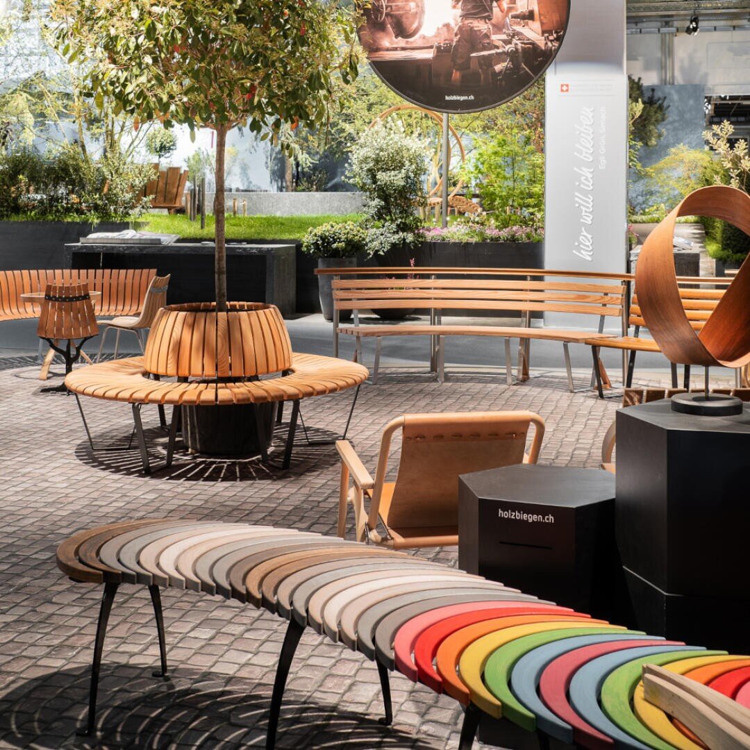 Eine moderne Outdoor-Möbelausstellung zeigt Holzbänke, eine kreisförmige Bank um einen Baum und eine wellenförmige, regenbogenfarbene Bank auf einer Kopfsteinpflasterfläche, umgeben von Pflanzen und Grünzeug.