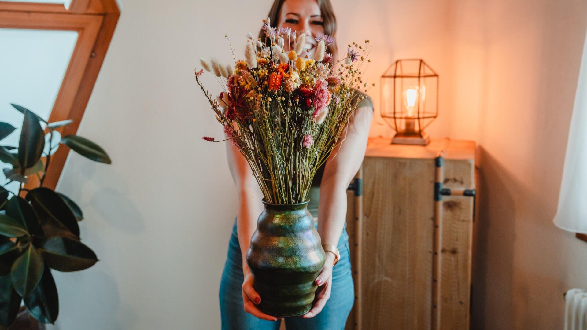 Eine Frau steht in einem Raum und hält eine große Vase mit Trockenblumen in die Kamera. Das warme Licht einer Lampe beleuchtet sanft den Raum, der gemütlich und rustikal eingerichtet ist.
