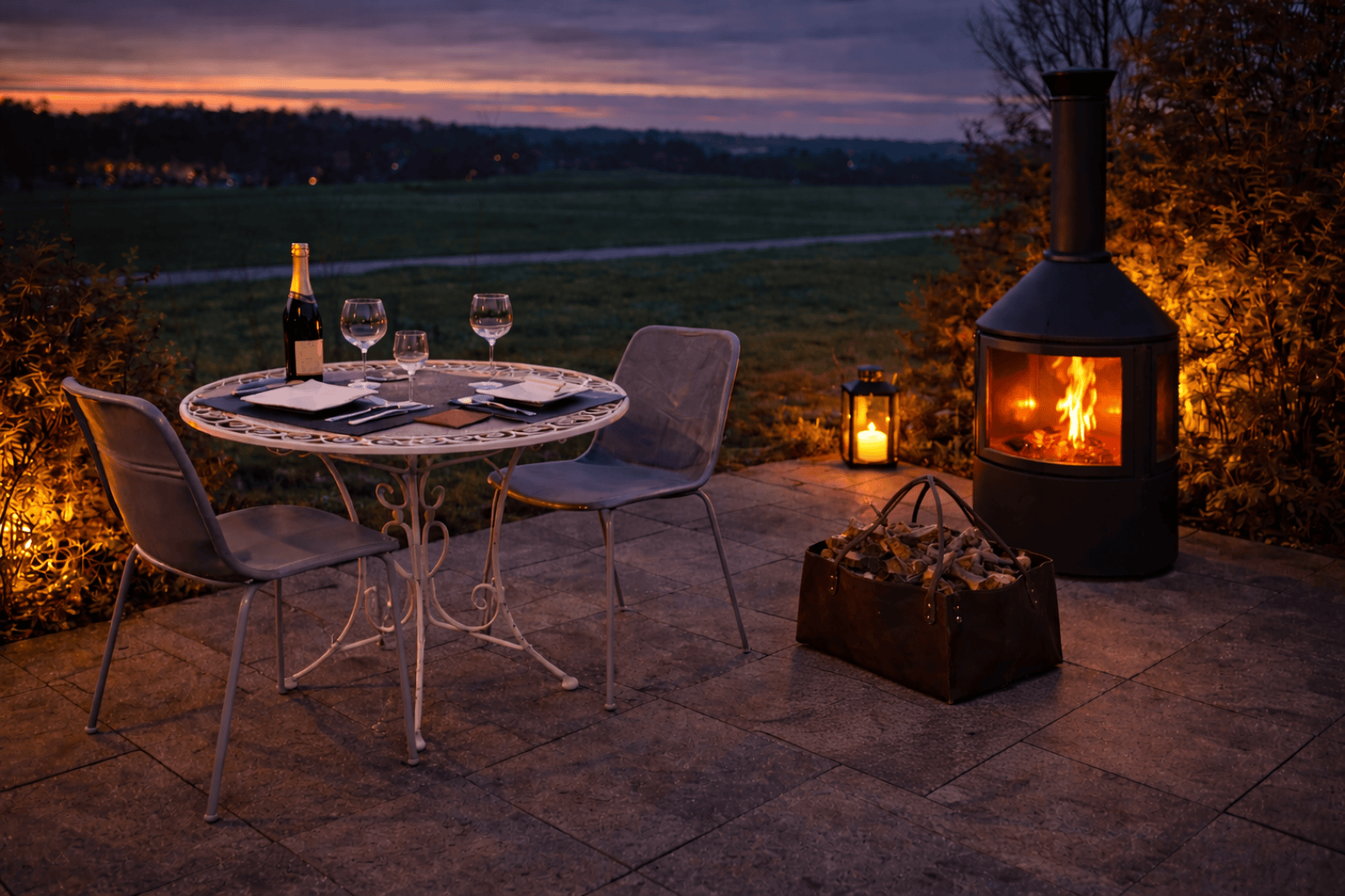 A cozy patio scene at sunset with a round table for two, wine glasses, a champagne bottle and notebooks. Two chairs stand in front of a lighted outdoor fireplace with lanterns and a basket of firewood nearby.
