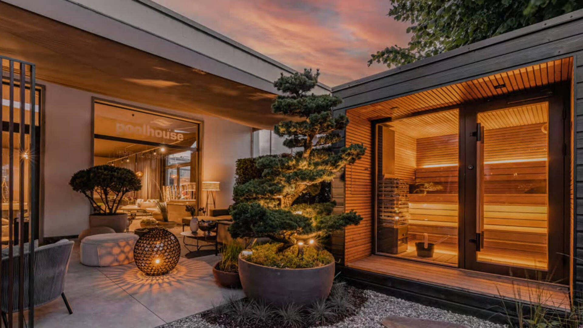 Ein moderner Außenbereich bei Sonnenuntergang mit einer warm beleuchteten Sauna, einem skulptierten Baum in einem großen Pflanzgefäß, gemütlichen Sitzgelegenheiten und einem Schild mit der Aufschrift Poolhouse in einem modernen Gebäude.