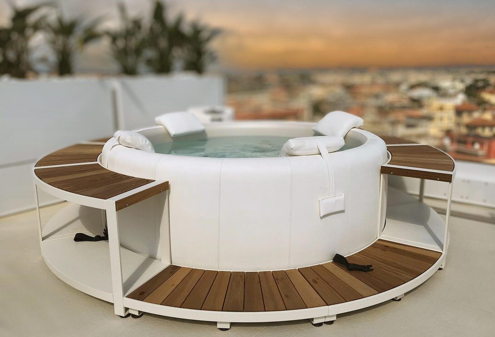 Auf einer Dachterrasse steht ein weißer, runder Whirlpool mit hölzernen Sitzflächen und Rückenlehnen. Im Hintergrund verschwimmt die Stadtkulisse, und am Rand sind Topfpflanzen zu sehen.