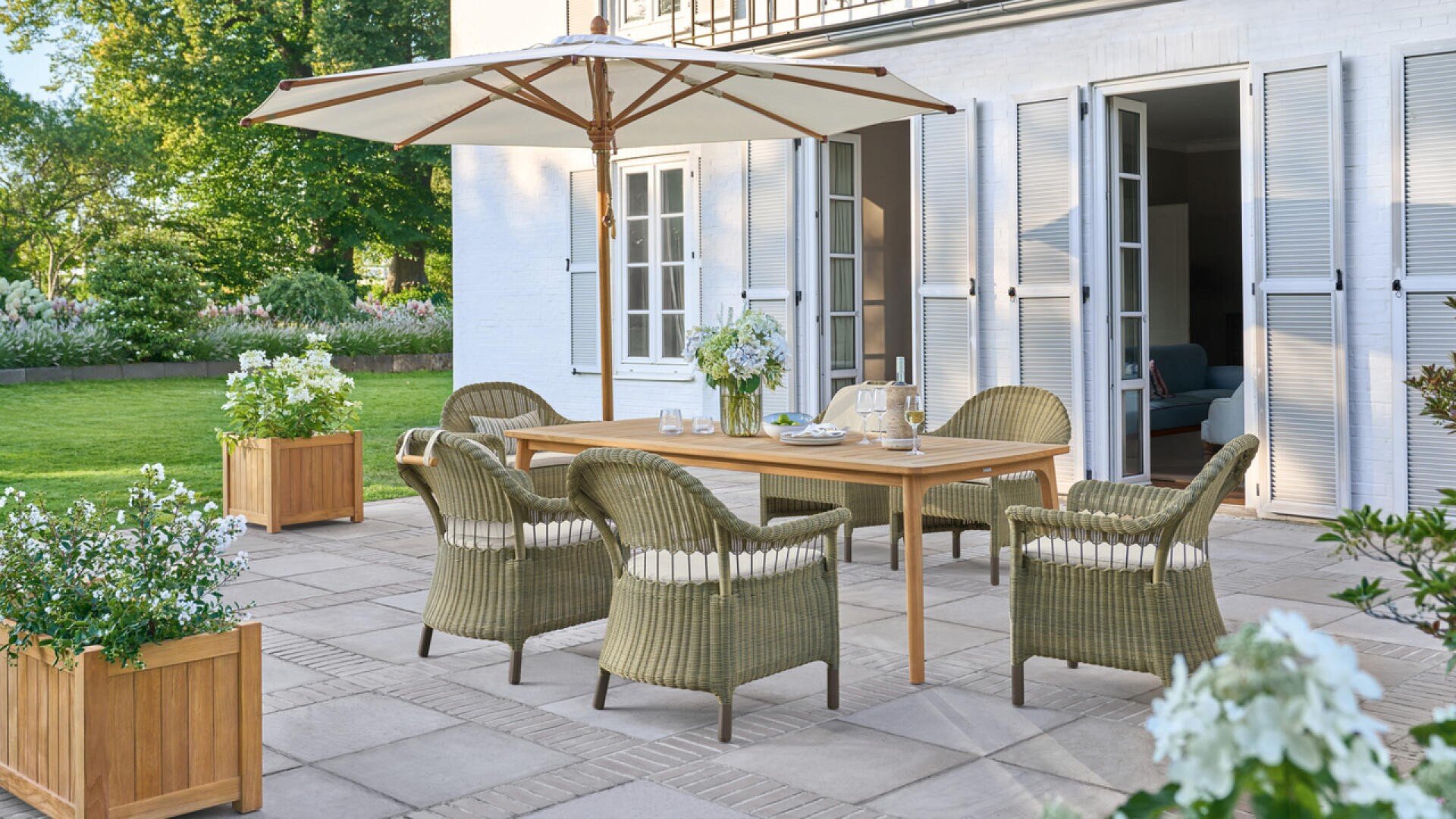 Eine Terrasse mit einem Holztisch, fünf Korbstühlen, einem großen weißen Sonnenschirm und Topfblumen, die an einem sonnigen Tag vor einem weißen Haus mit offenen Flügeltüren steht und von Grün umgeben ist.