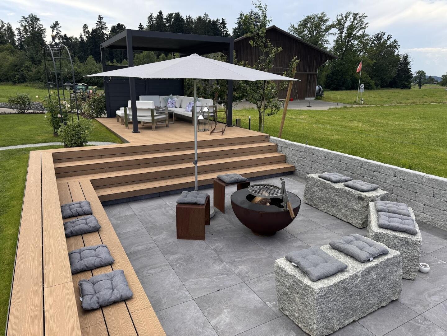 Moderne Außenterrasse mit gepolsterten Steinbänken, einer Feuerstelle und einem großen Sonnenschirm. Eine Holztreppe führt zu einem überdachten Lounge-Bereich mit Sofas. Der Platz ist von Gras und Bäumen umgeben.
