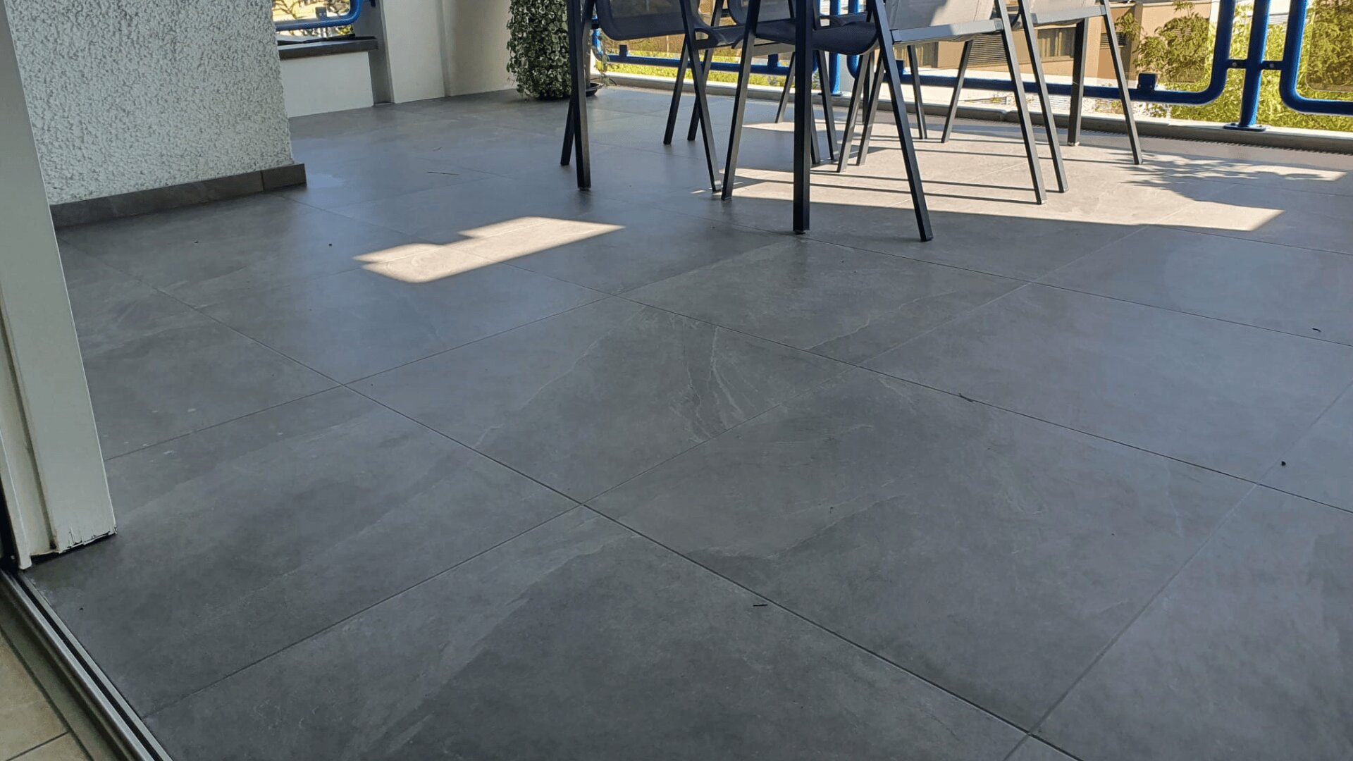 Geräumige Terrasse mit großen grauen Bodenfliesen, Teilansicht eines Tisches und Stühlen, Glasbalkongeländer und Grünpflanzen, die an einem sonnigen Tag draußen sichtbar sind.