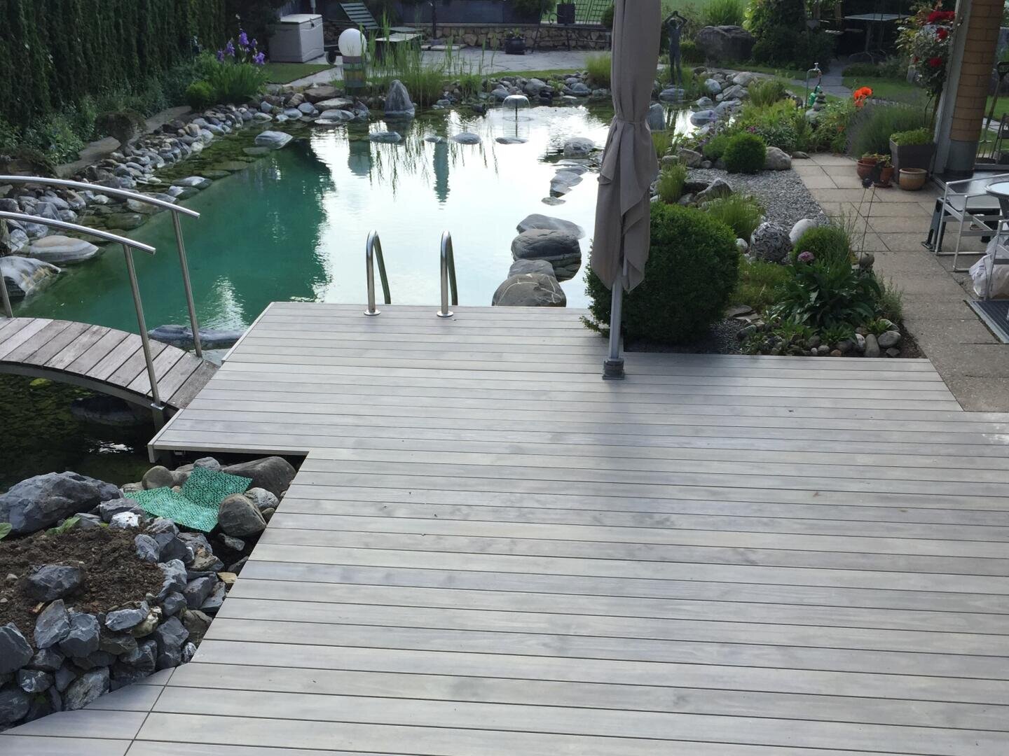 Eine Holzterrasse führt zu einem natürlichen Schwimmteich mit klarem Wasser, der von Steinen, Grünpflanzen und Terrassenmöbeln umgeben ist. Der Zugang ist über eine Metallleiter möglich und eine kleine Brücke führt über einen Teil des Teichs.