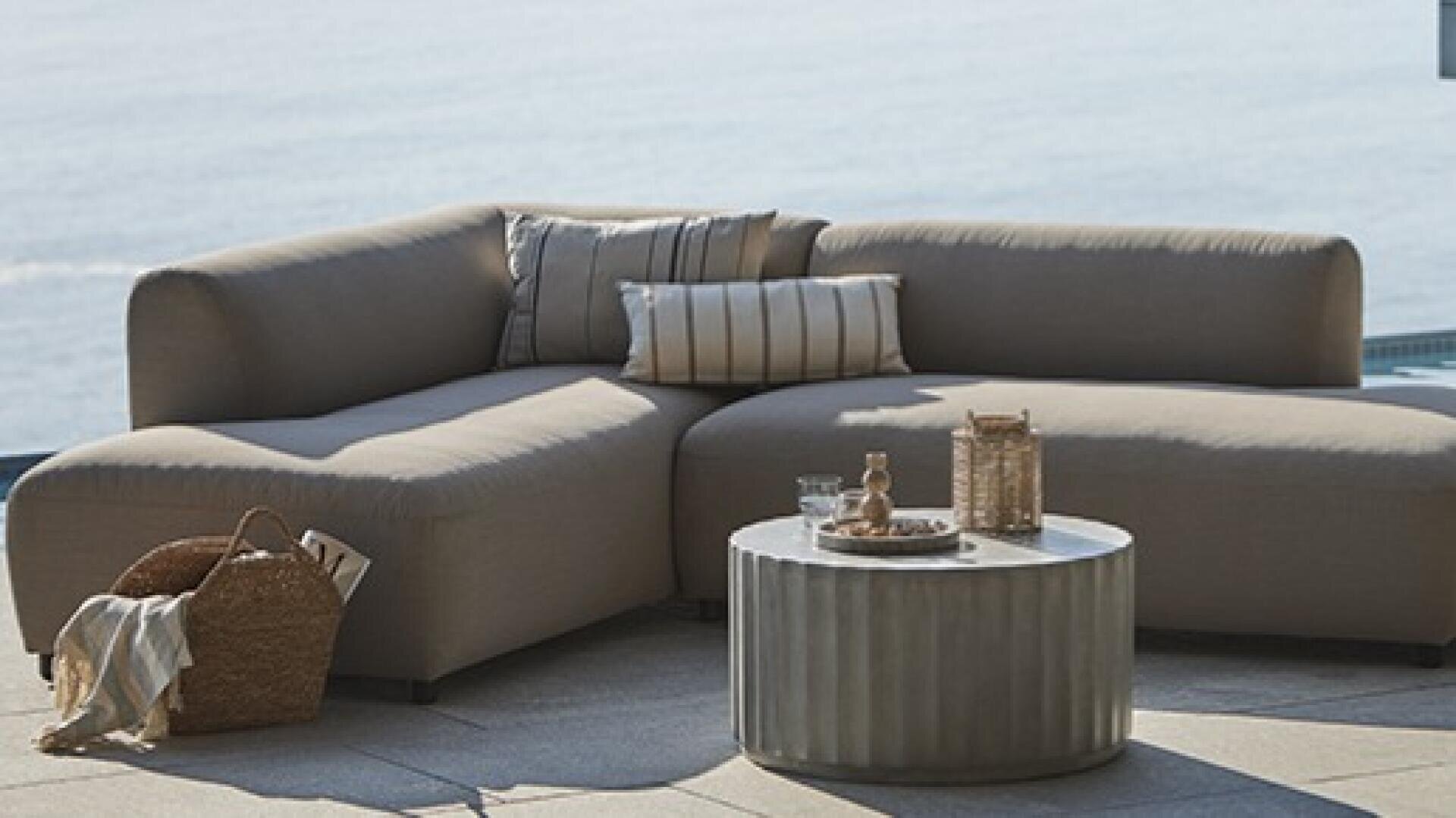 Ein modernes Outdoor-Sectional-Sofa mit beigen Kissen und gestreiften Wurfkissen sitzt neben einem runden Couchtisch mit geflochtenen Dekorationsartikeln und blickt auf einen ruhigen Meerblick. In der Nähe steht ein geflochtener Korb mit einer Decke auf dem Boden.