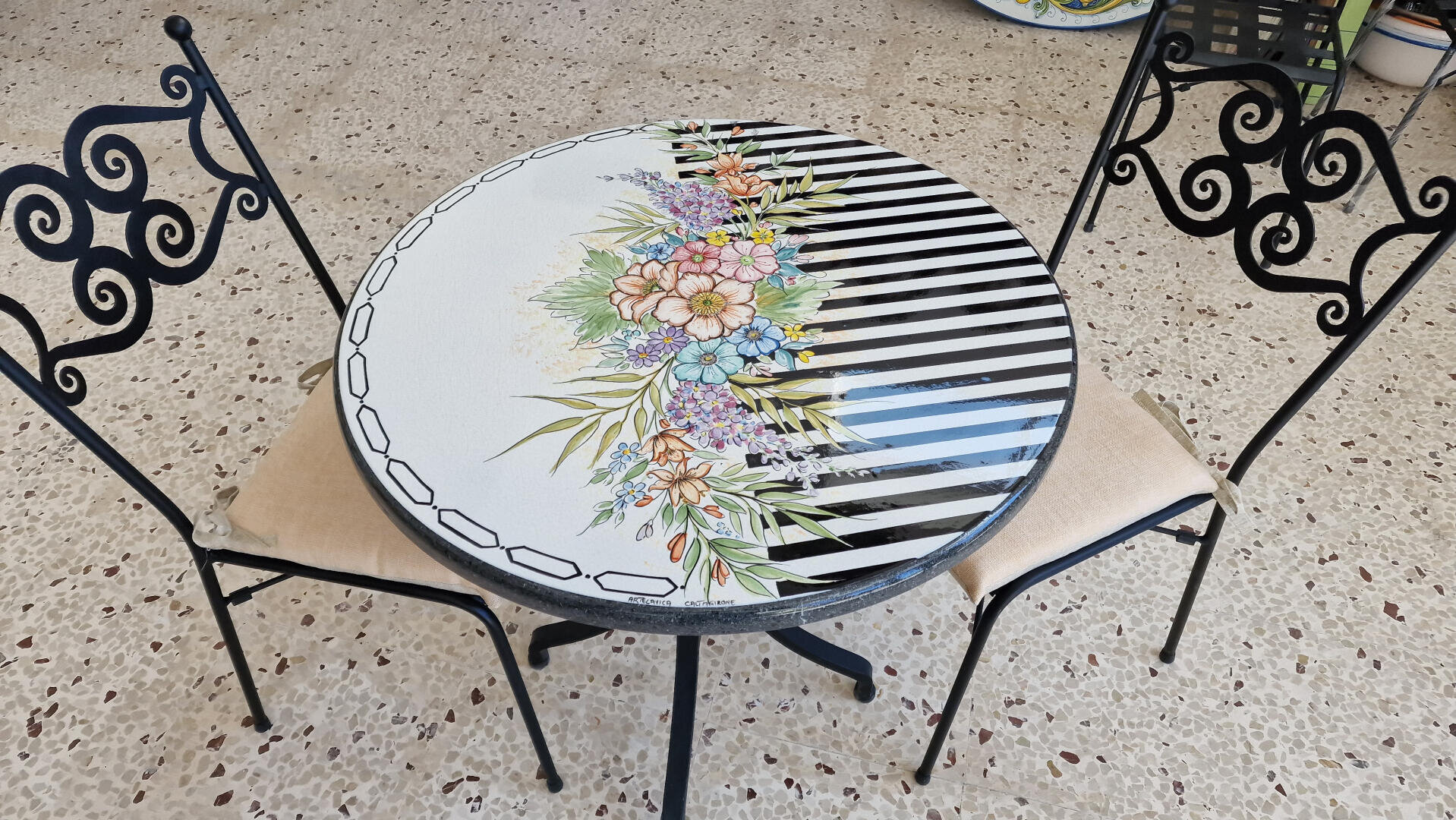 A round table with a floral and black-striped design on the surface is surrounded by two black metal chairs with beige cushions on a terrazzo floor.