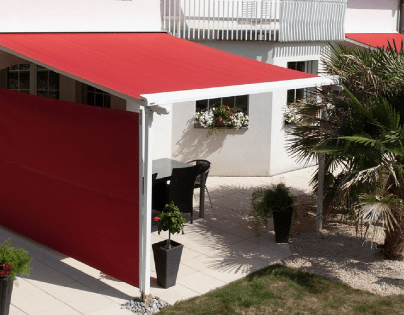 Eine Terrasse mit roter Markise und passendem roten Seitenschirm, Tisch und Stühlen im Freien, Topfpflanzen und einer Palme, angrenzend an ein weißes Gebäude mit Blumenkästen am Fenster.