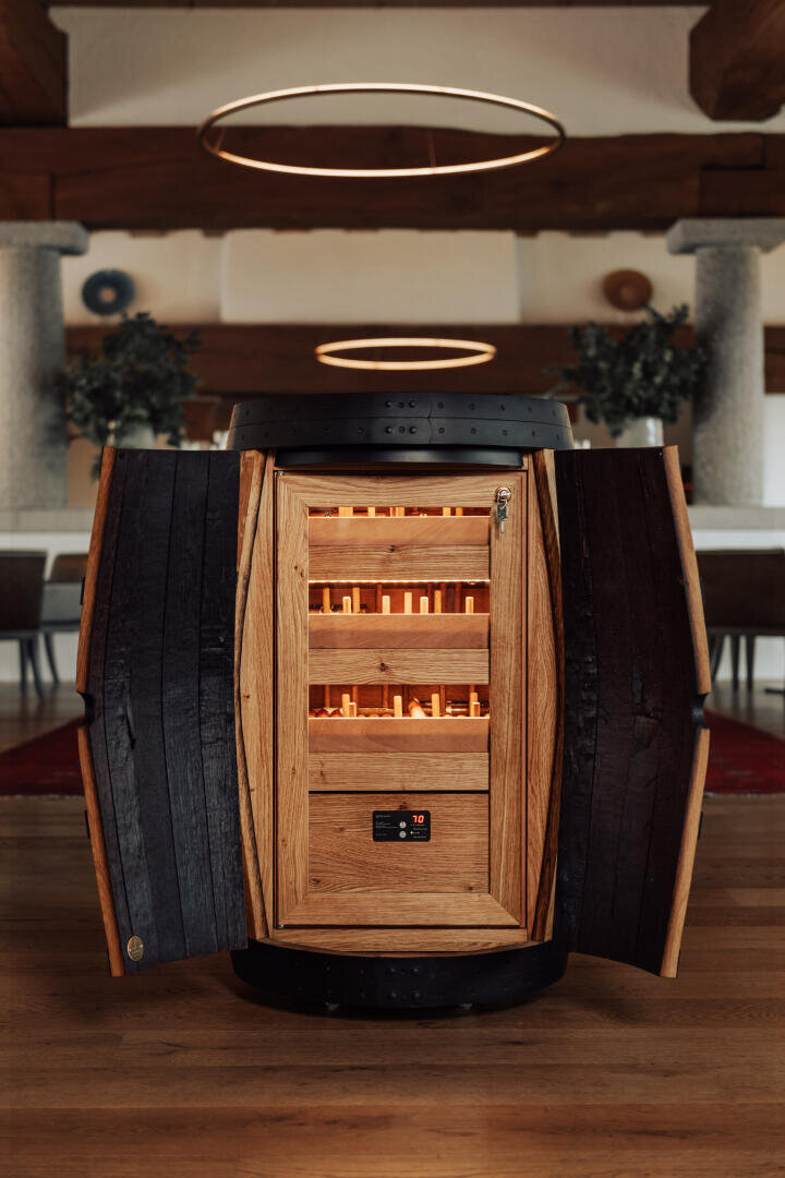 Ein als Humidor umfunktioniertes Holzfass steht in einem modernen Raum, dessen Türen sich öffnen und den Blick auf geordnete Zigarrenregale freigeben, die von der Innenbeleuchtung sanft beleuchtet werden.