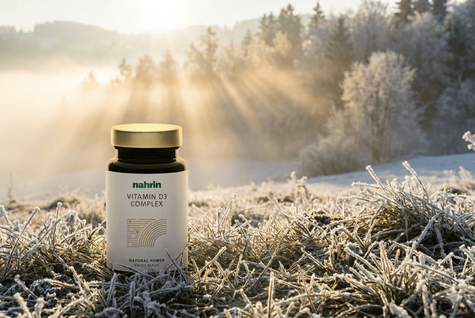 A bottle of nahrin Vitamin D3 Complex stands on a frosty grass in a sunny, foggy outdoor setting with sunlight filtering through the trees in the background.