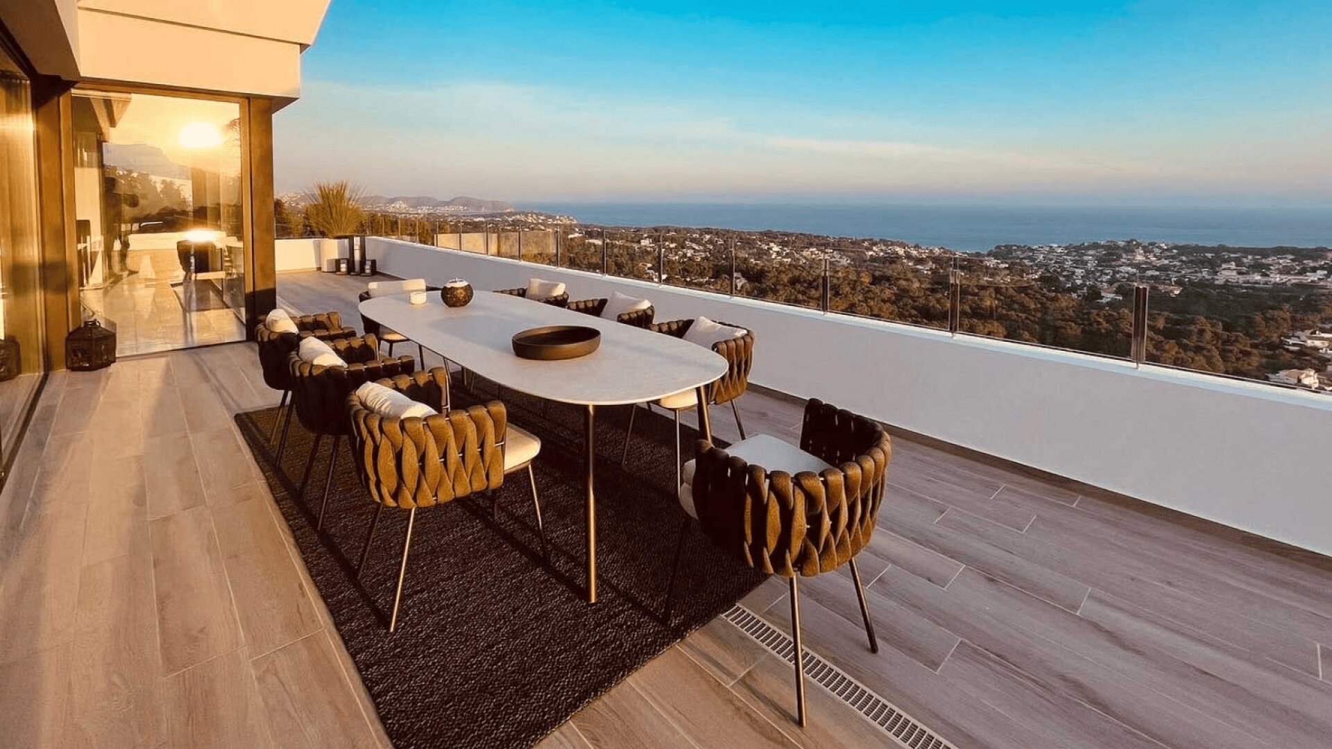 Moderner Essbereich im Freien auf einem Balkon mit Korbstühlen und einem langen Tisch, mit Blick auf eine malerische Aussicht auf Hügel, Häuser und das Meer bei Sonnenuntergang.