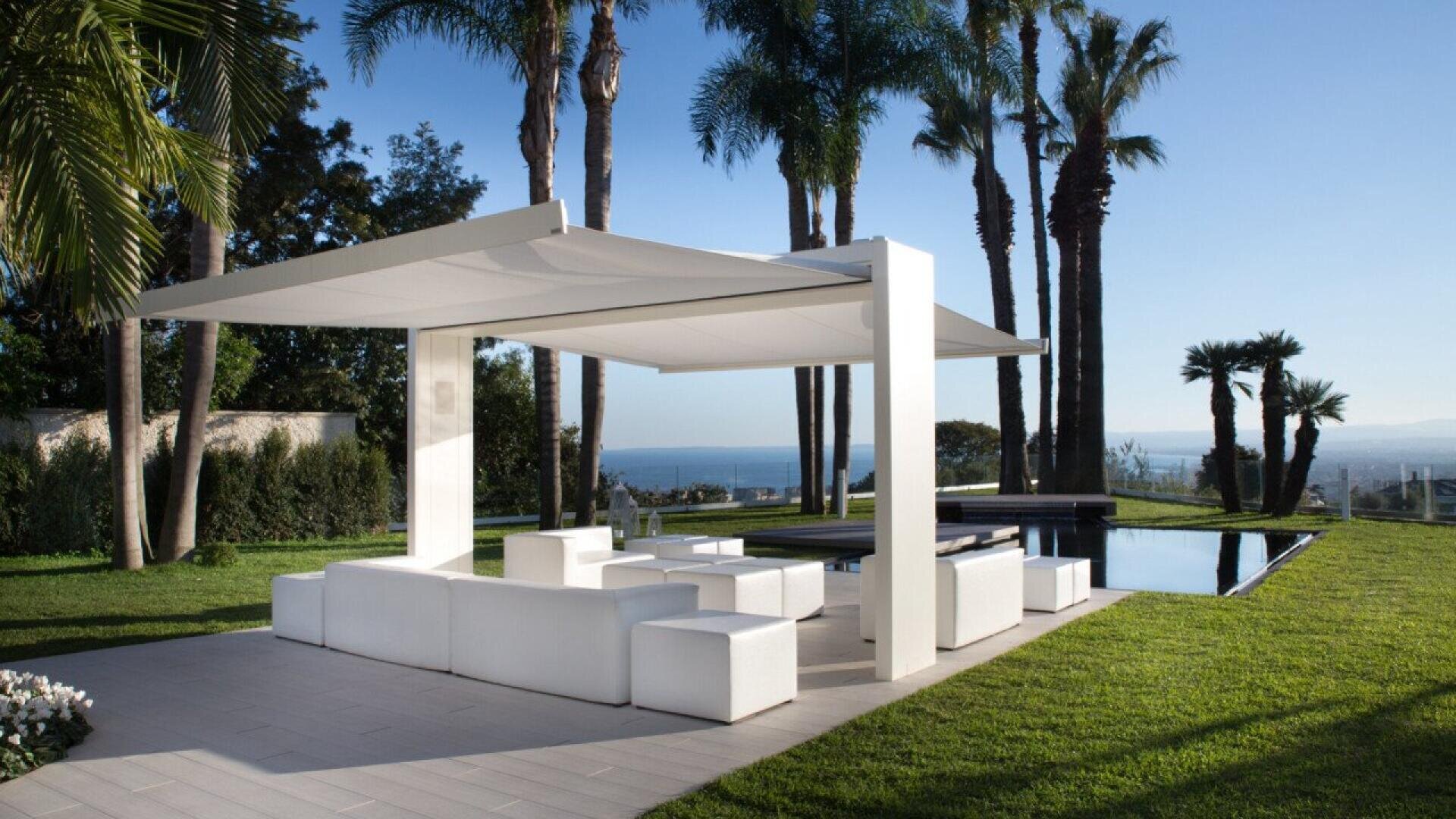 A modern outdoor lounge area with white seating and a canopy is set beside a rectangular pool, surrounded by green grass, palm trees, and overlooking a scenic distant view under a clear blue sky.