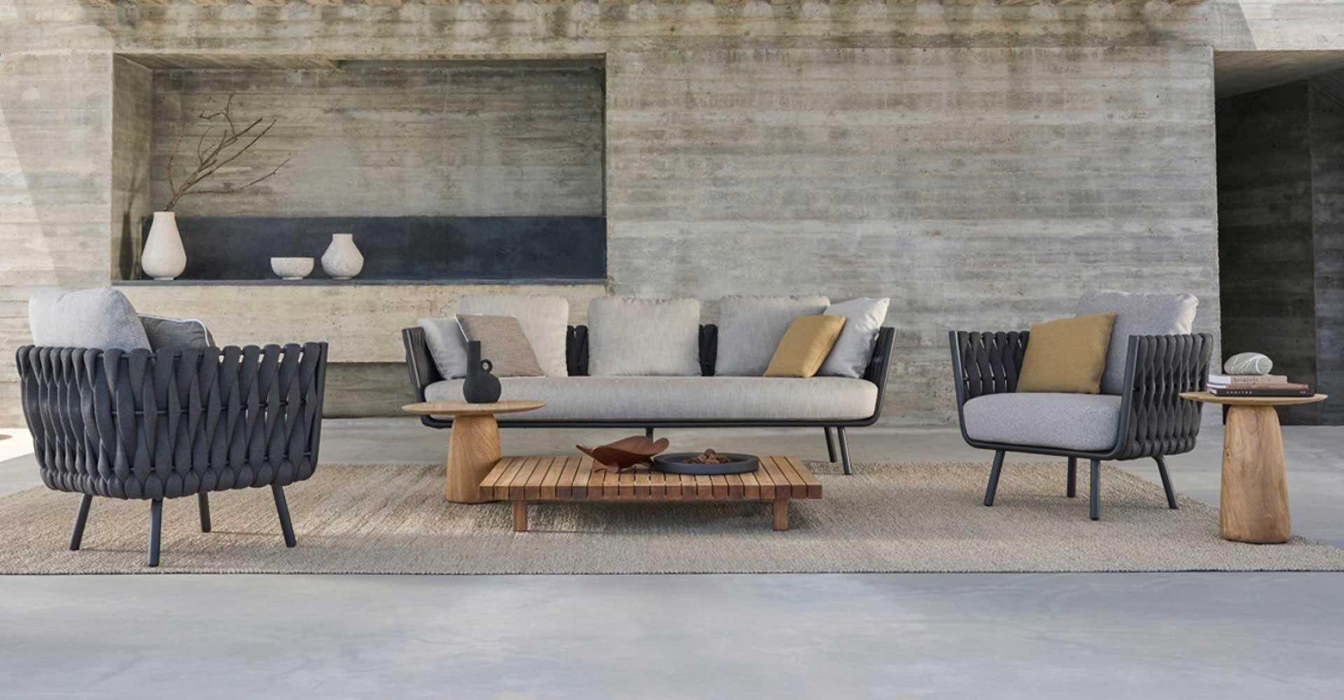 Modern outdoor seating area with a neutral-toned sofa and two armchairs with a woven frame, wooden side tables, a low coffee table and minimalist vases in front of a concrete wall.
