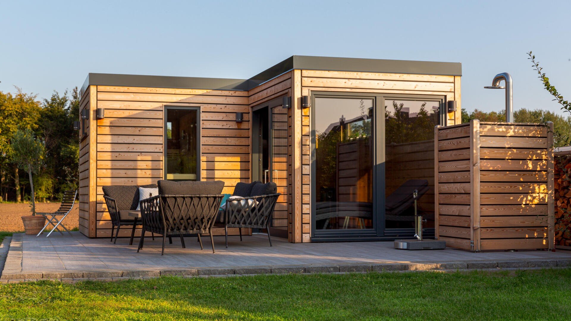 Ein modernes kleines Haus mit Holzverkleidung verfügt über eine Terrasse mit Sitzgelegenheiten im Freien und einem Tisch. In der Nähe befindet sich eine Außendusche mit Sichtschutzwänden, und das Haus ist von grünem Gras und Bäumen umgeben.