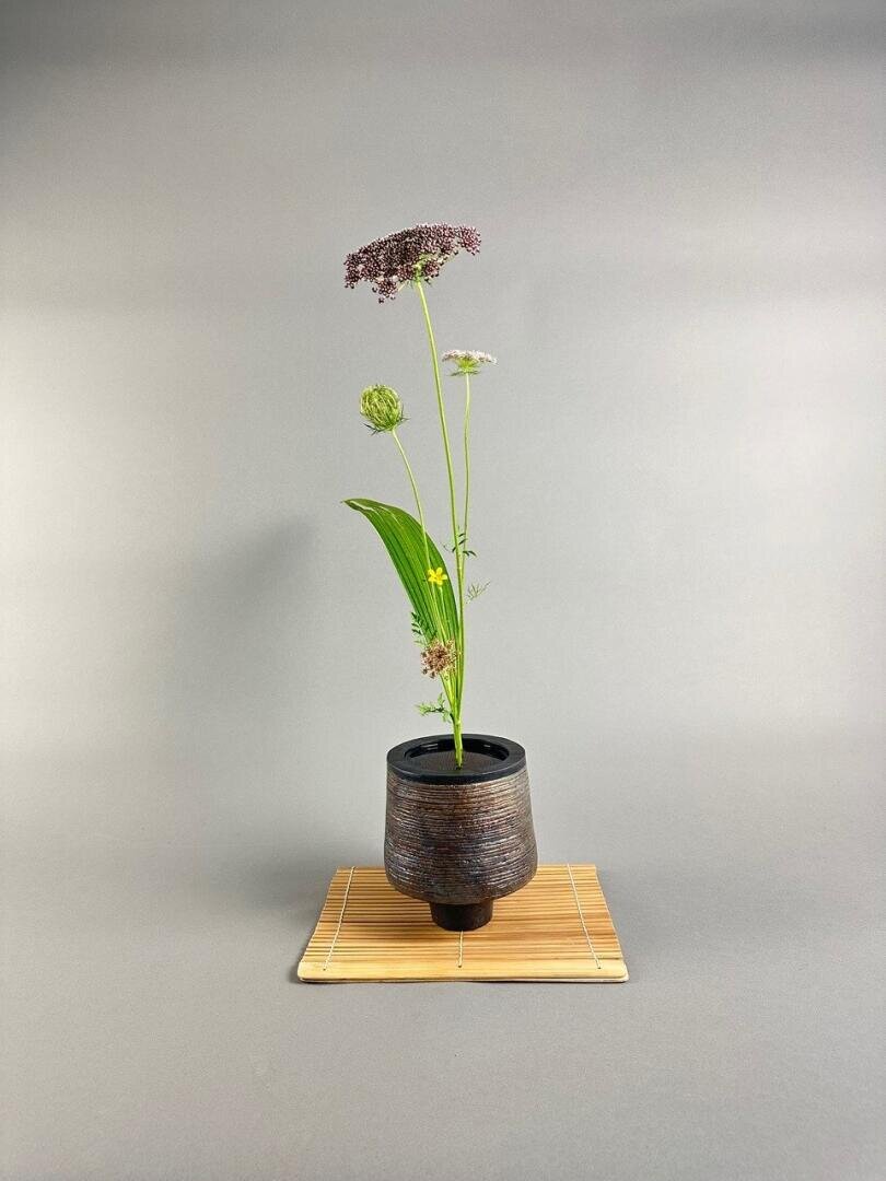 Ein minimalistisches Blumenarrangement mit ein paar hohen grünen Stängeln und zarten lila und weißen Blumen in einer dunklen Keramikvase, die auf einer Bambusmatte vor einem schlichten grauen Hintergrund platziert ist.