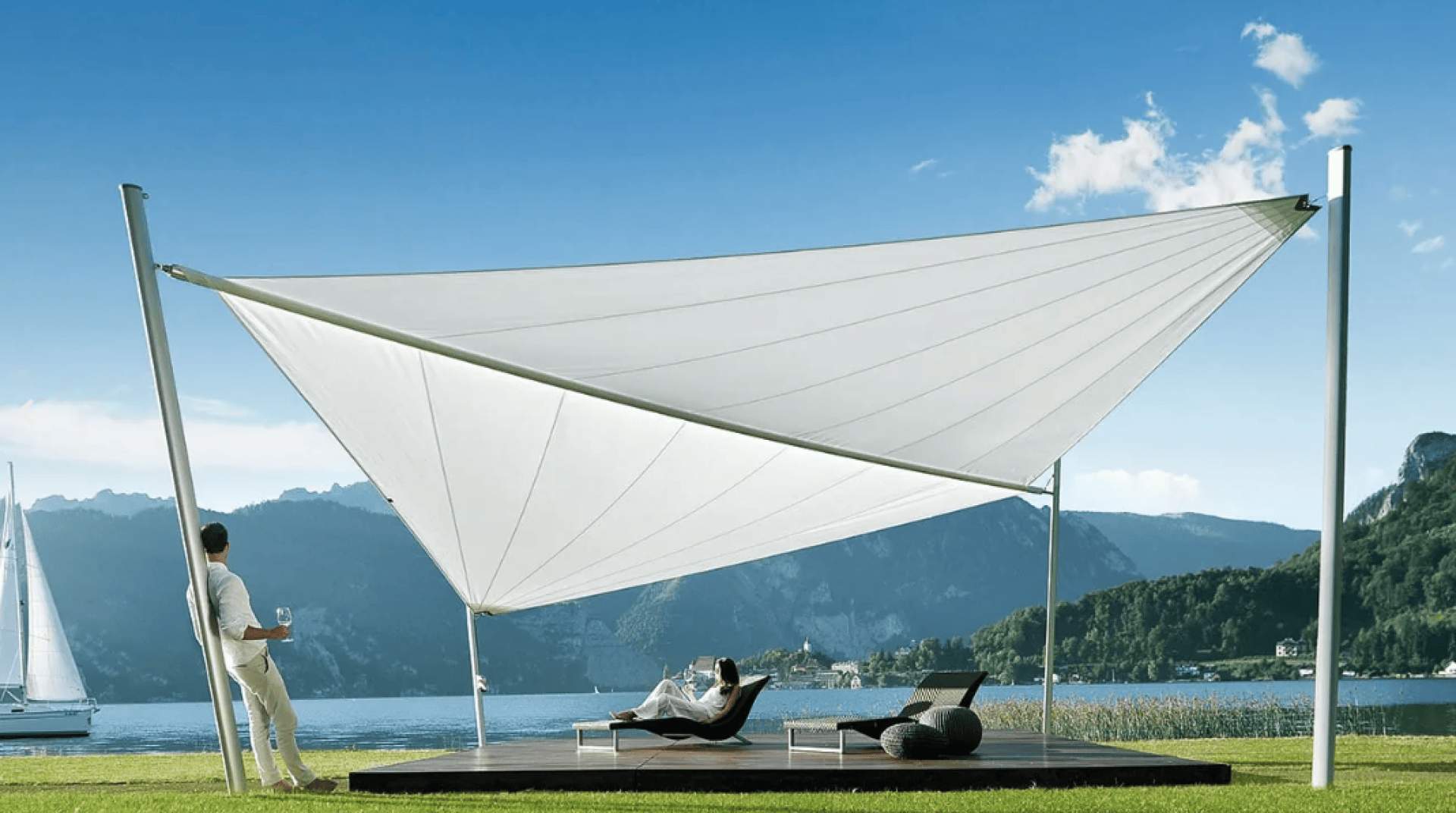 A person stands holding a drink while another relaxes on a lounge chair under a modern white canopy by a lake, with mountains and a sailboat in the background on a clear sunny day.
