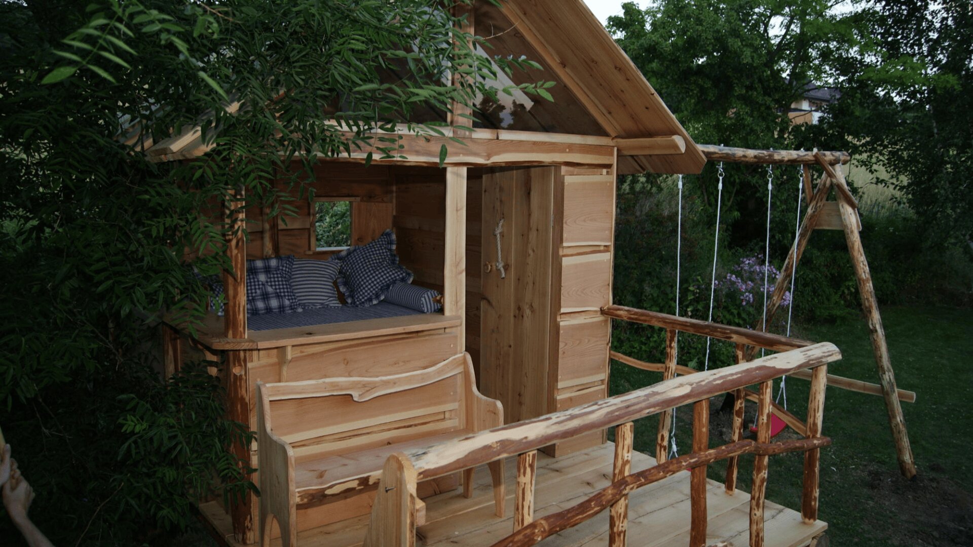 Ein hölzernes Baumhaus mit einem offenen Fenster, einer kleinen Terrasse mit einer Bank und einer Schaukel. Im Inneren des Baumhauses sind Kissen zu sehen, und der Bereich ist von grünen Bäumen und Gras umgeben.