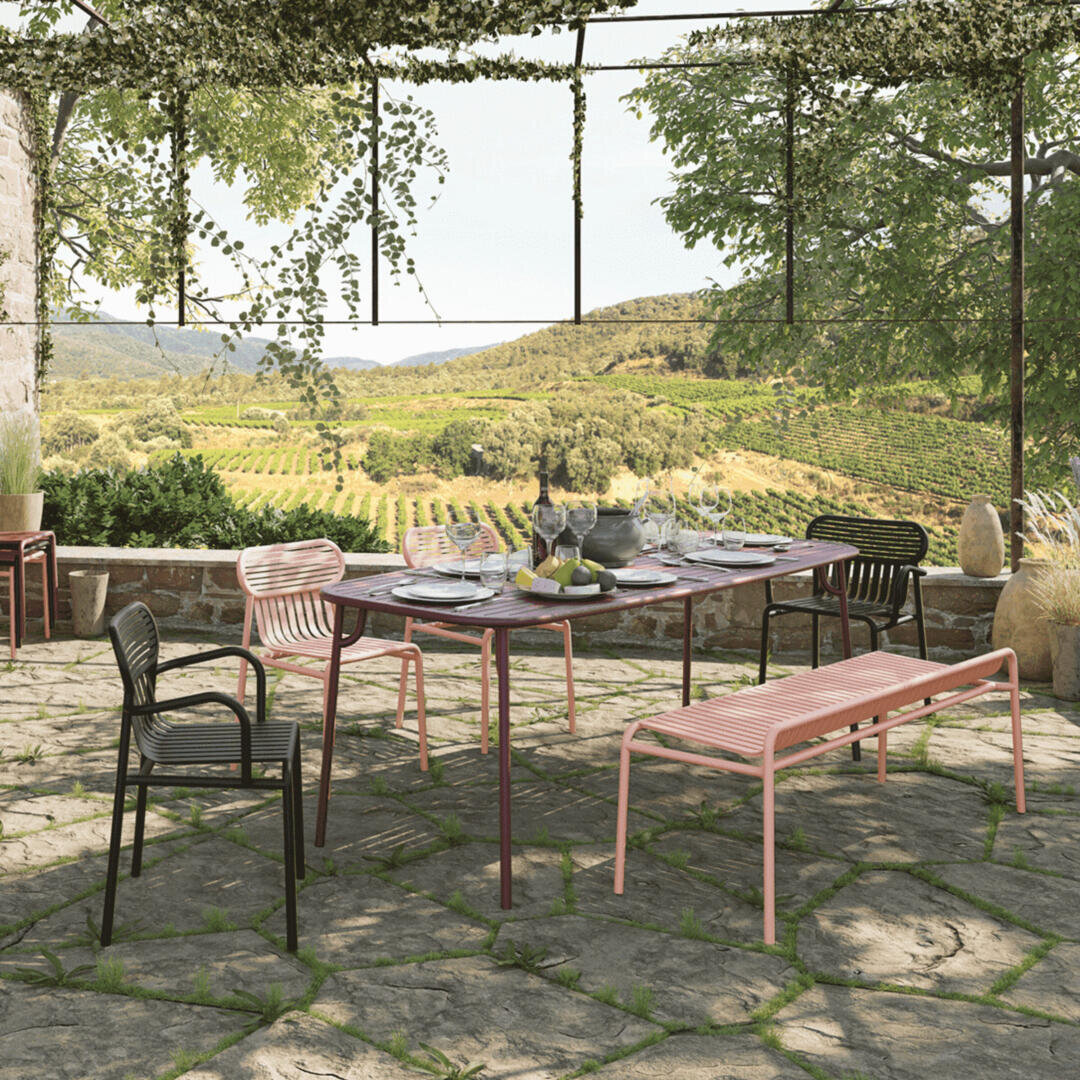 Essbereich im Freien mit einem für eine Mahlzeit gedeckten Tisch, umgeben von modernen Stühlen und Bänken auf einer Steinterrasse. Im Hintergrund sind üppige Weinberge und sanfte Hügel zu sehen, unter einer Pergola mit Weinreben.