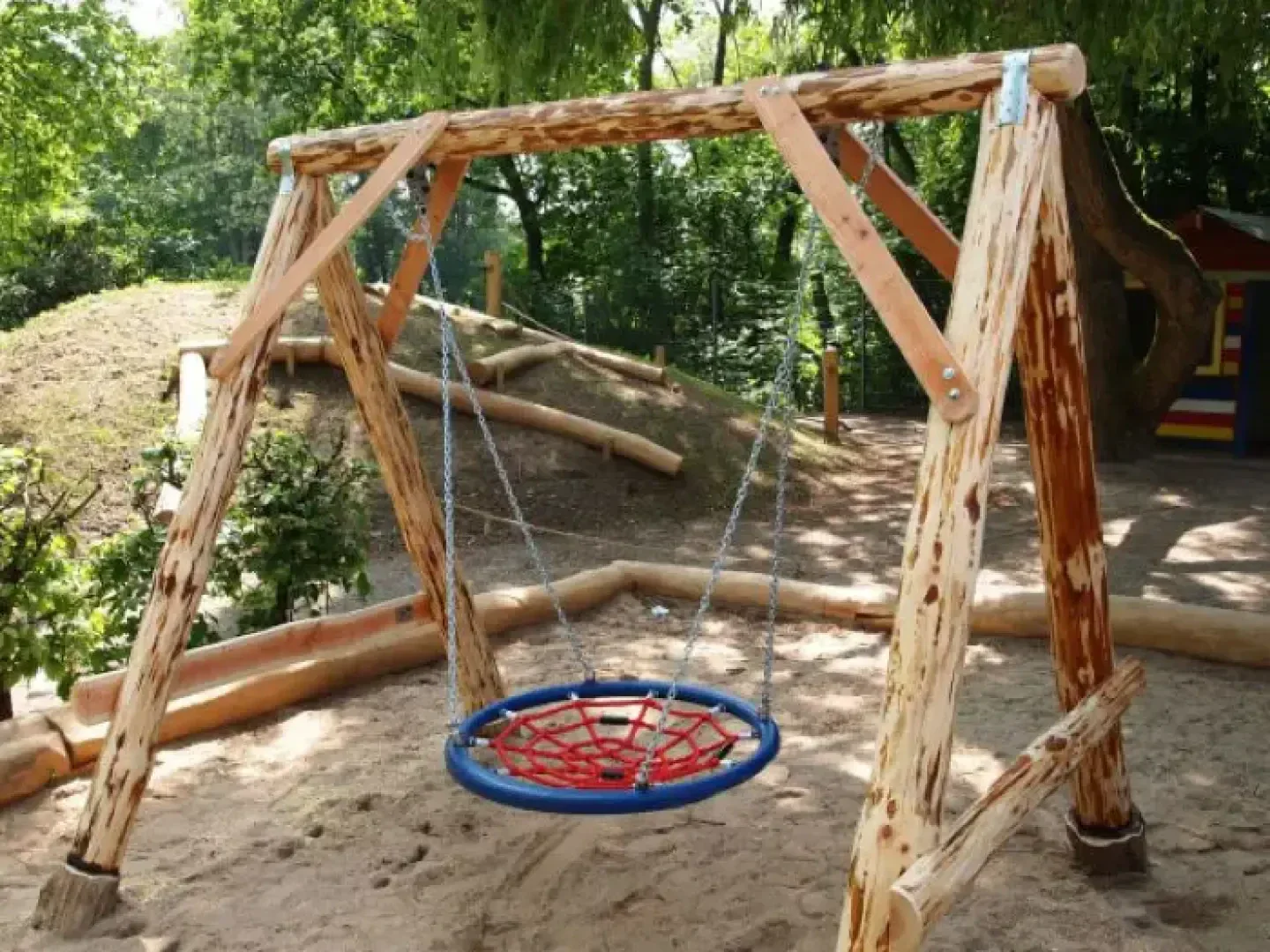 A wooden swing set with a round, netted blue and red swing hangs over a sandy area in a sunny, green playground surrounded by trees and wooden borders.