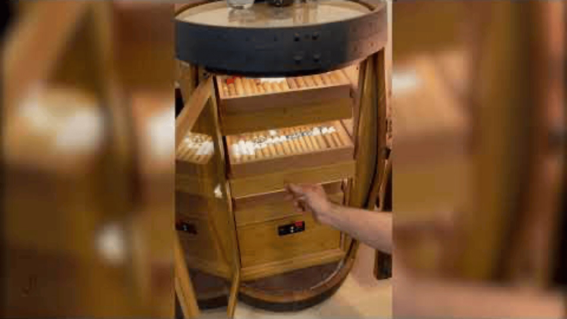 Eine Hand zieht eine mit Zigarren gefüllte Schublade in einem runden Holzhumidor mit mehreren Regalen auf, in denen die Zigarren fein säuberlich angeordnet sind. Der Humidor hat Glastüren und digitale Bedienelemente.