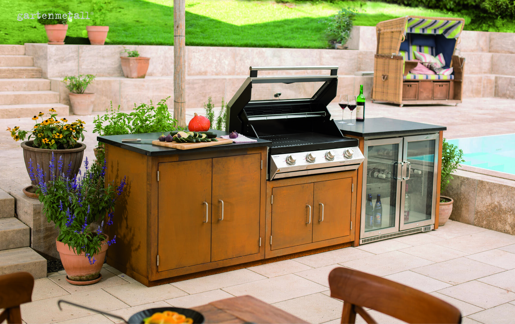Die Außenküche besteht aus einem Edelstahlgrill mit geöffnetem Deckel, einer Arbeitsplatte mit Gemüse und einem Schneidebrett, einem Mini-Kühlschrank mit Glasfront, zwei Gläsern Rotwein und Topfpflanzen in der Nähe eines Pools.