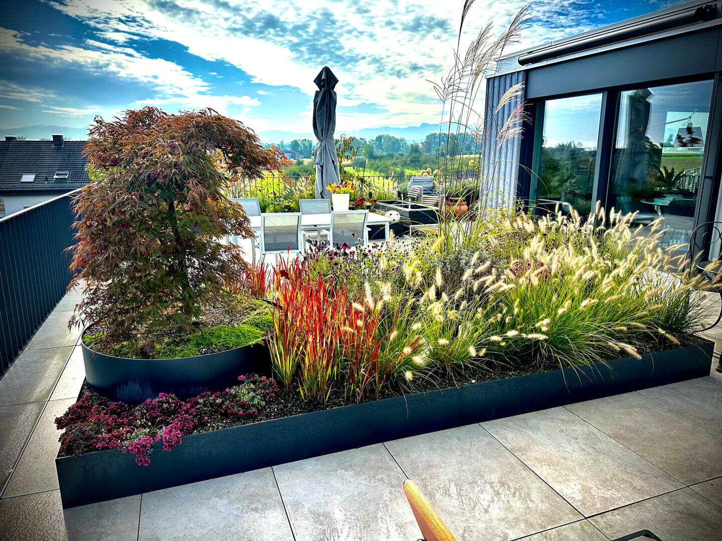 A modern rooftop garden with ornamental grasses, small trees, and colorful plants in raised beds, next to an outdoor dining area with white chairs and a large closed umbrella under a blue sky.