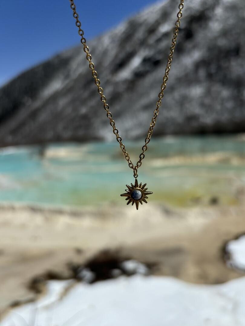 Eine goldene Halskette mit einem sonnenförmigen Anhänger mit einem kleinen Edelstein in der Mitte wird im Freien gehalten, mit einem verschwommenen Hintergrund aus türkisfarbenem Wasser und schneebedeckten Bergen.
