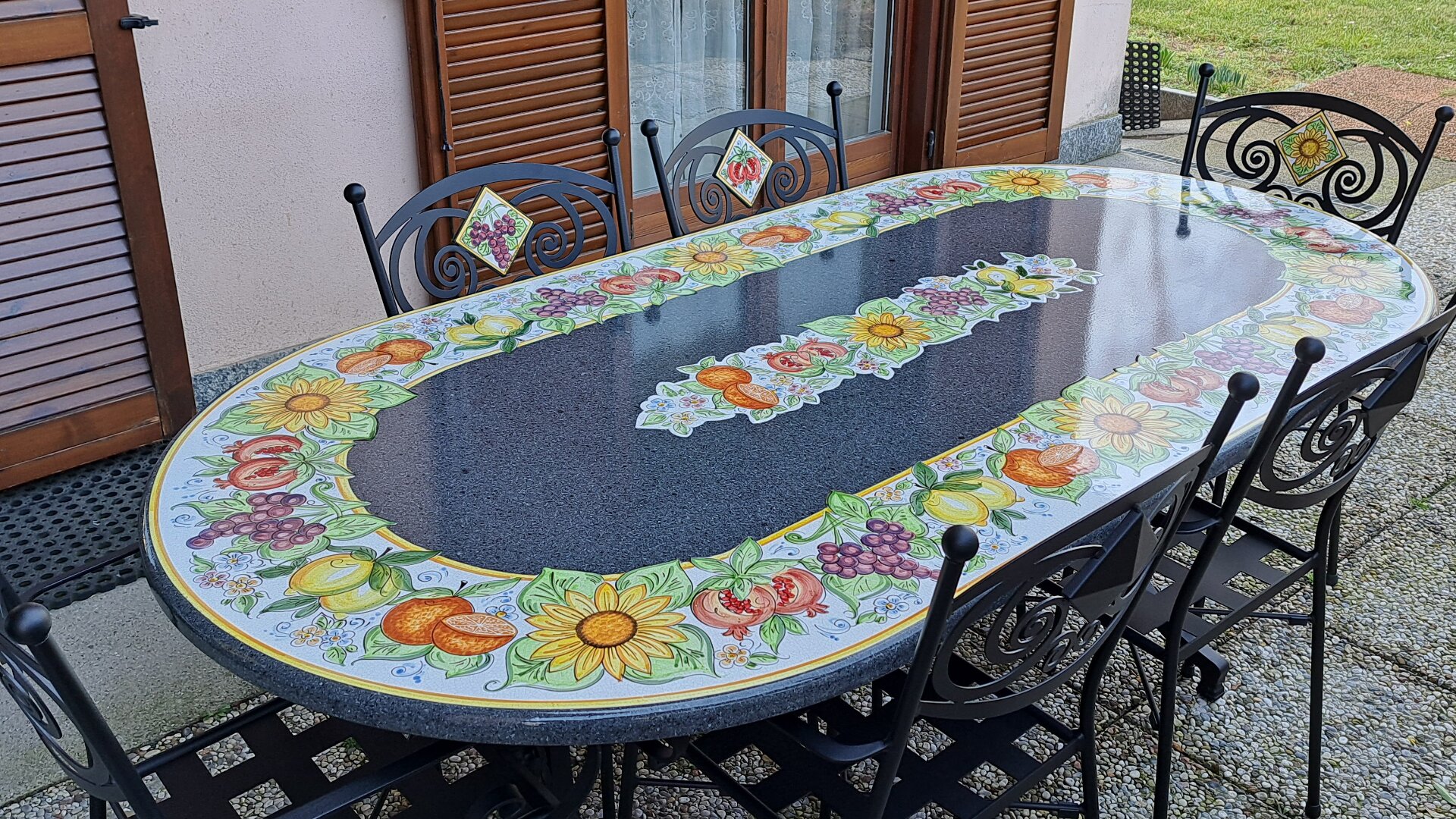 Ein großer ovaler Tisch für den Außenbereich mit einem glänzend schwarzen Zentrum und einer bunten Umrandung mit gemalten Sonnenblumen, Früchten und Blättern, umgeben von sechs schwarzen Metallstühlen mit dekorativen Rückenlehnen.