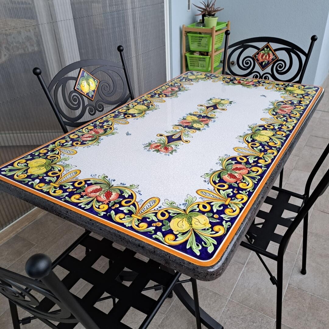 Ein rechteckiger Tisch mit einer bunten, floral gemusterten Keramikplatte, umgeben von drei schwarzen Metallstühlen mit dekorativen Rückenlehnen, steht auf einem gefliesten Balkon oder einer Terrasse. Im Hintergrund sind grüne Pflanzenregale zu sehen.