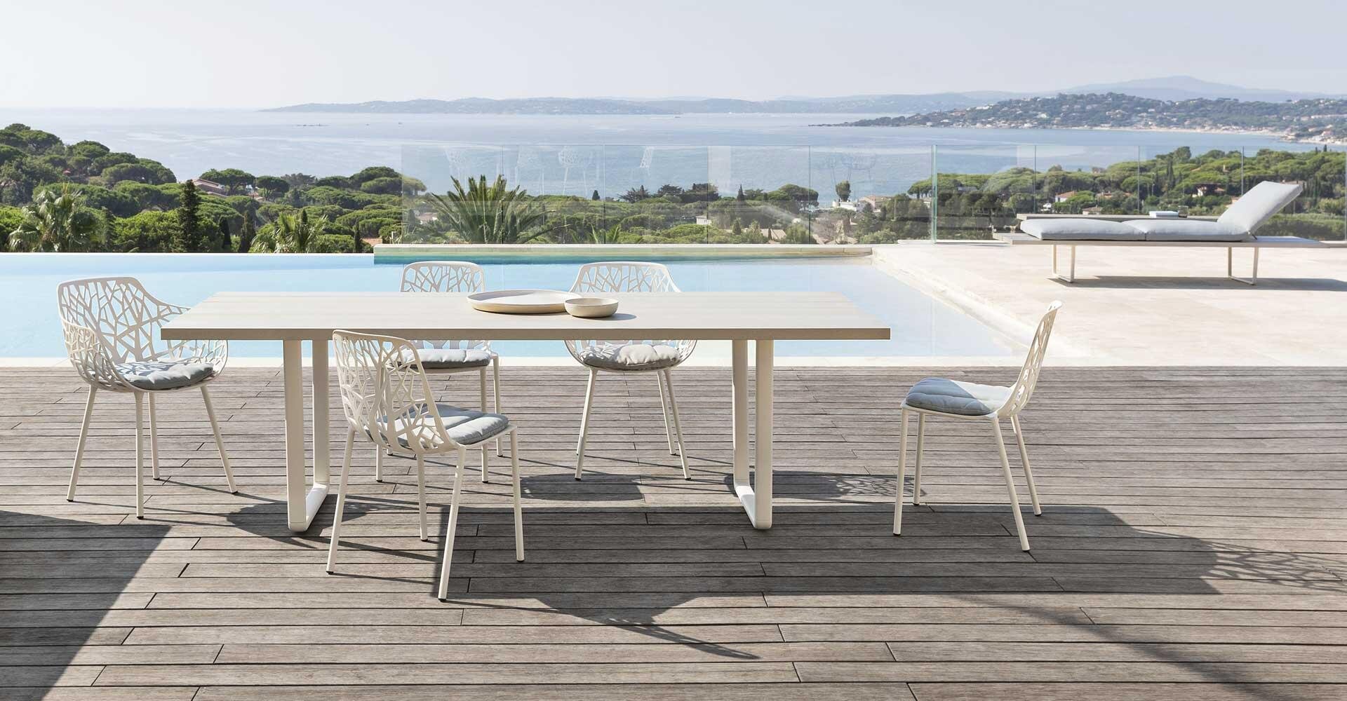 Modern outdoor dining room with a rectangular table and five white chairs on a wooden terrace next to a pool, overlooking a picturesque coastal landscape with trees, hills and calm water in the background.
