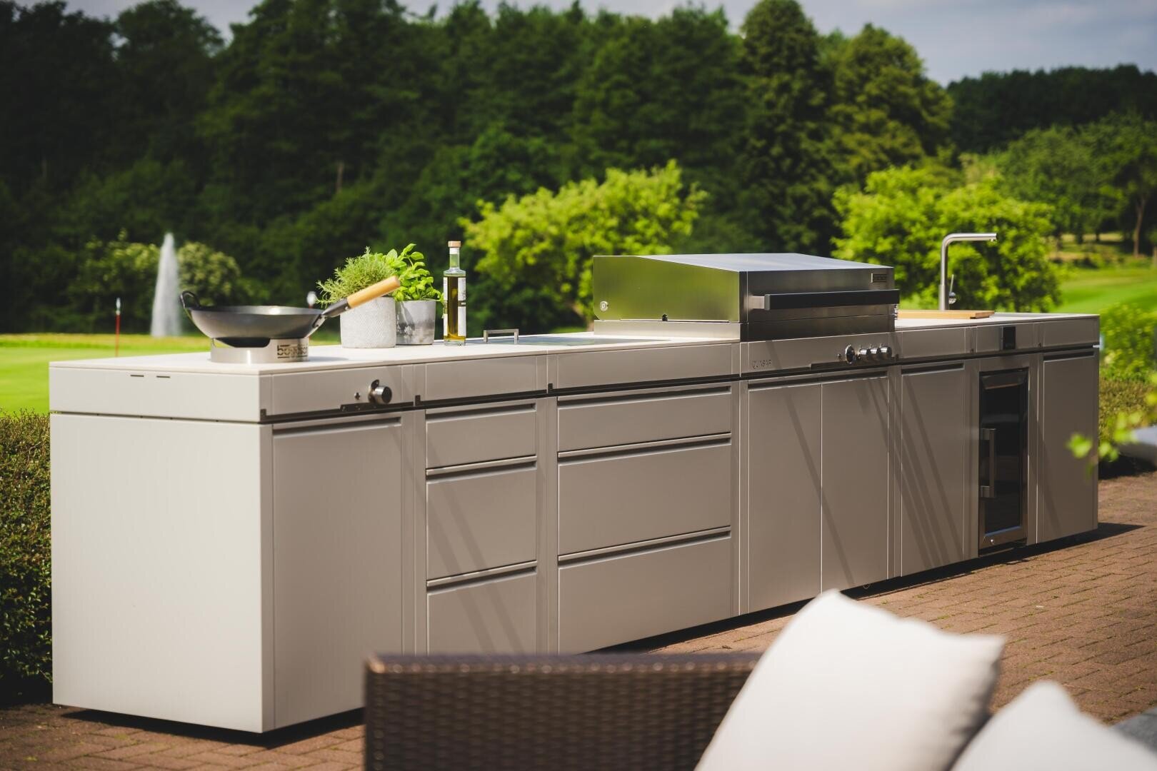 A modern outdoor kitchen with stainless steel cabinets, a built-in grill, sink, and cooking utensils, set on a patio with green trees and a fountain in the background.