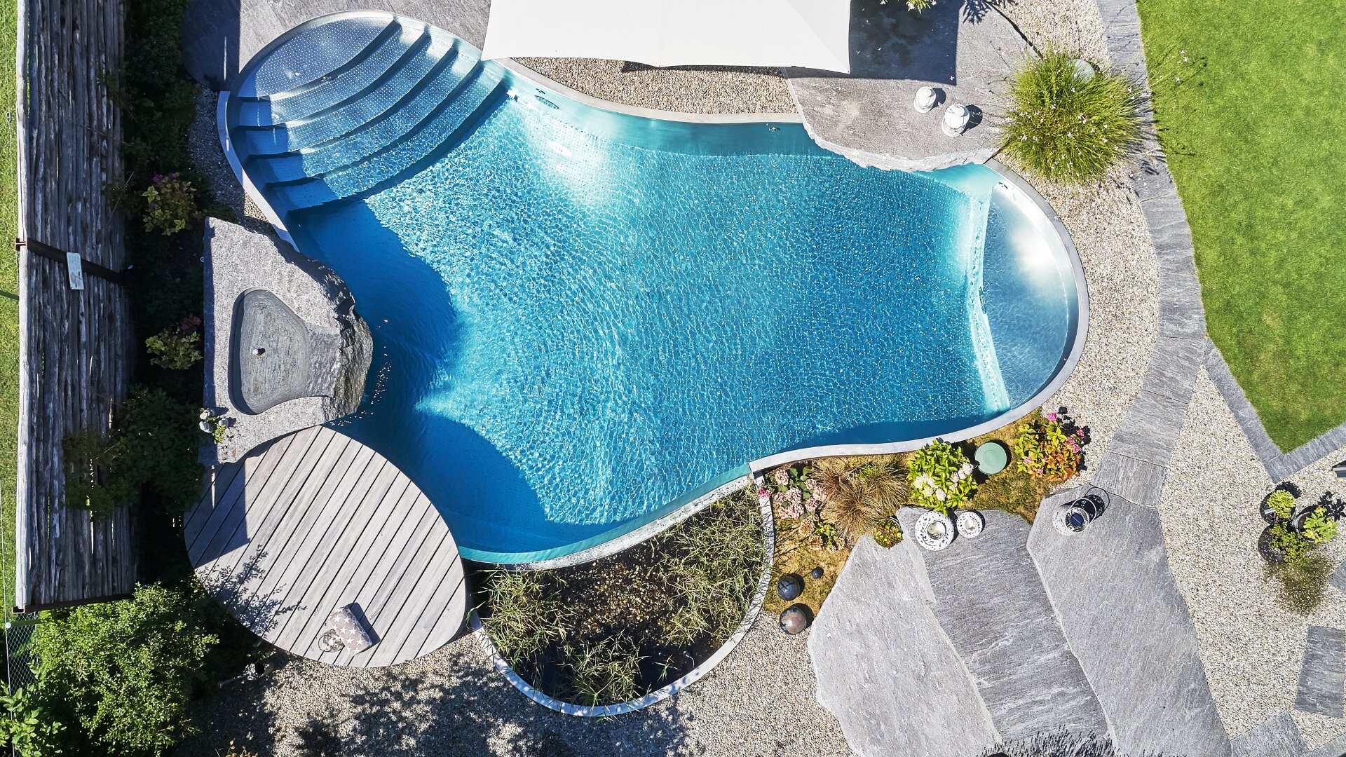 Luftaufnahme eines einzigartig geformten Außenpools mit geschwungenen Rändern, umgeben von Steinwegen, Kies, grünem Gras, Pflanzen und einem runden Holzdeck in der Nähe eines kleinen Whirlpools.