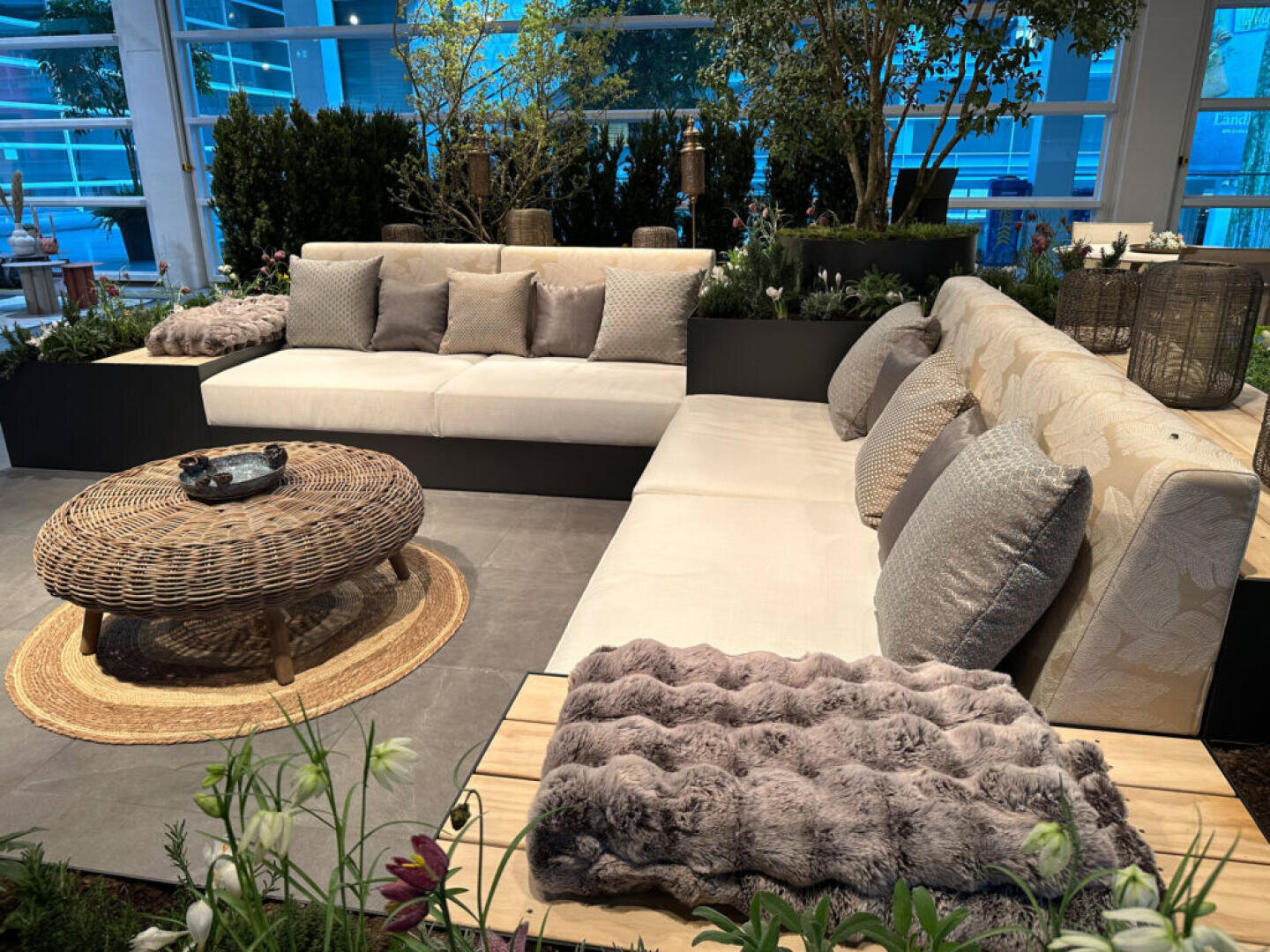 Modern outdoor seating area with two cream sofas, gray and beige cushions, a round wicker coffee table on a woven rug, lush green plants, and natural decor, set in a bright, glass-enclosed space.