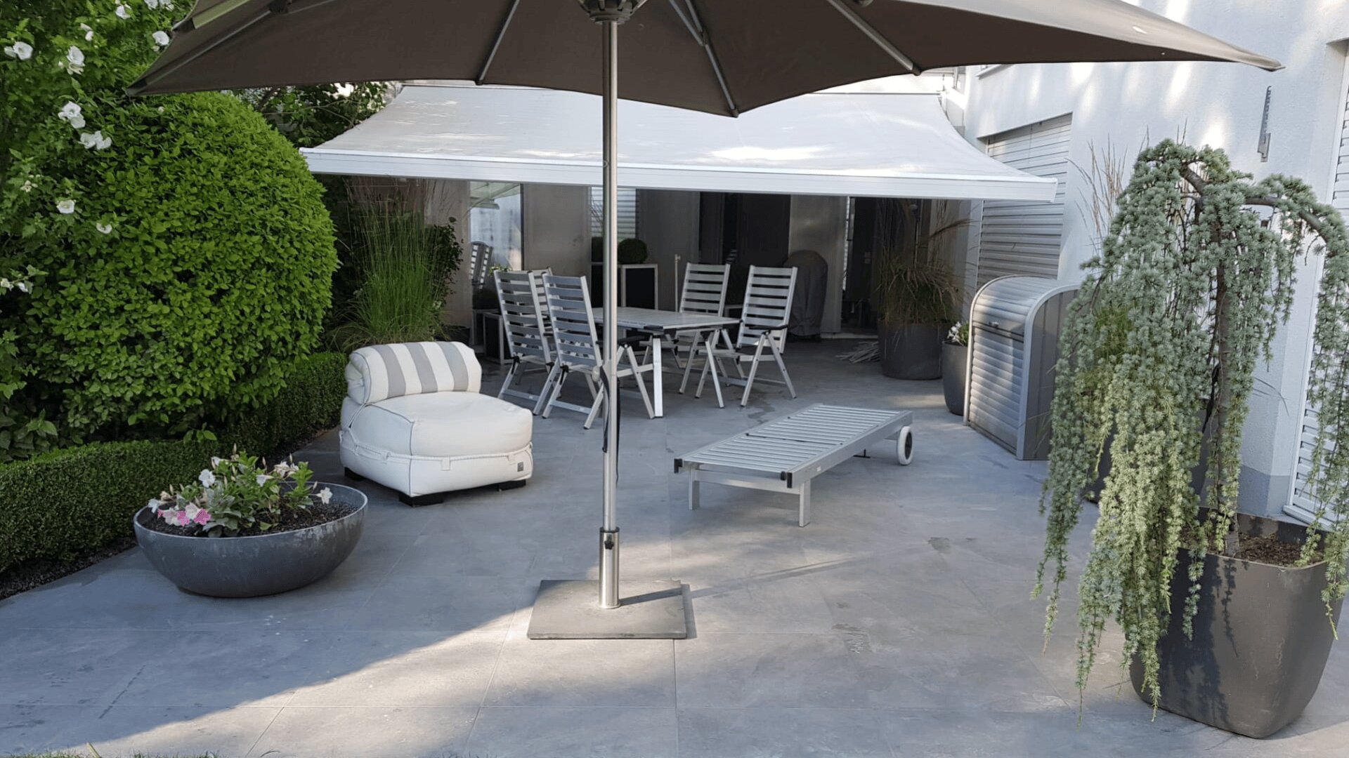 Auf einer modernen Terrasse mit grauen Fliesen stehen ein großer Sonnenschirm, ein weißer Loungesessel, eine weiße Sonnenliege, ein Esstisch mit Stühlen, Topfpflanzen und üppiges Grün an den Rändern. Ein weißes Vordach spendet zusätzlichen Schatten.