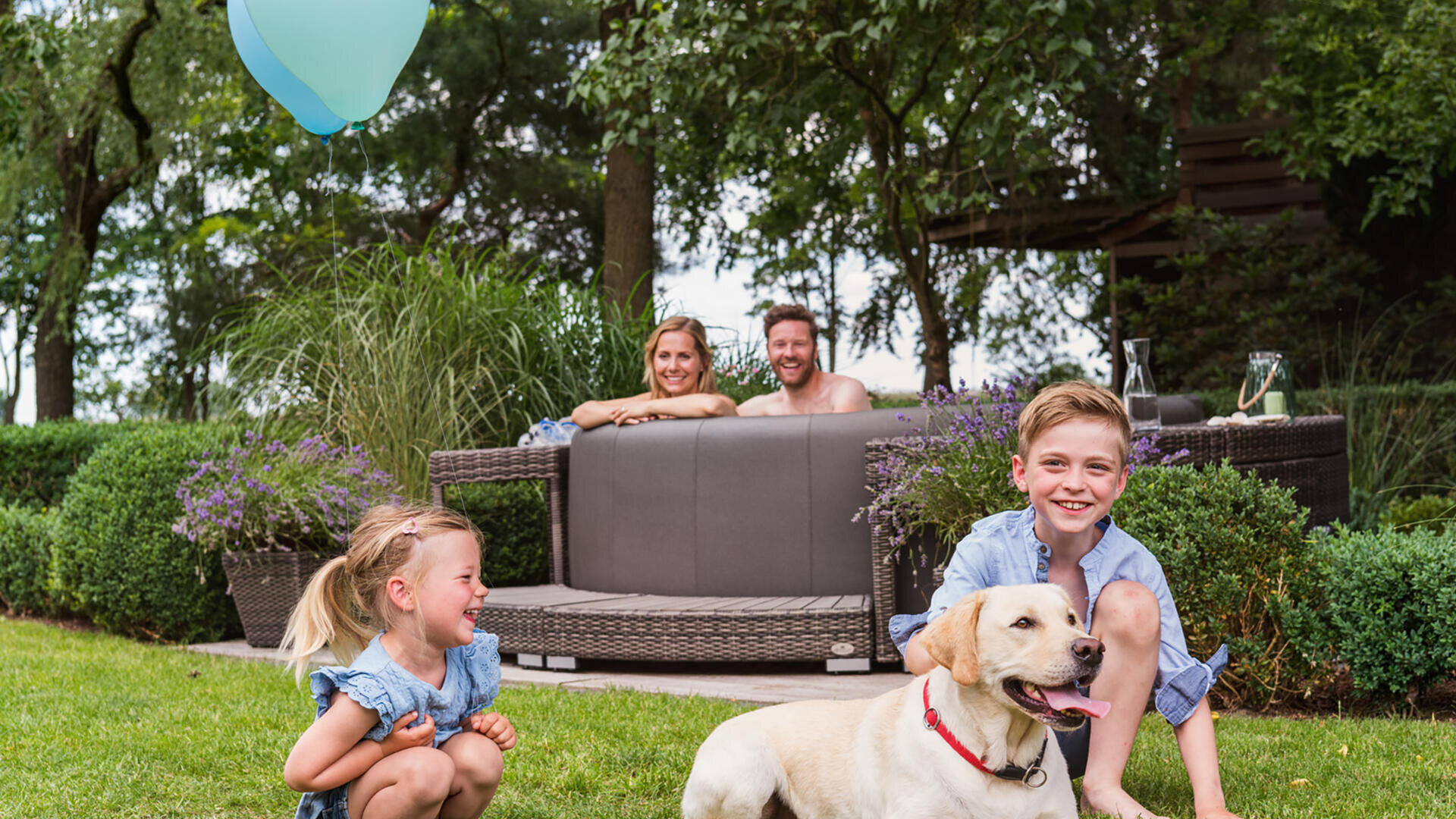 Zwei kleine Kinder spielen im Gras mit einem gelben Labradorhund; in der Nähe schwebt ein blauer Luftballon. Im Hintergrund sitzen zwei Erwachsene lächelnd auf einem Sofa im Freien, umgeben von Grün.