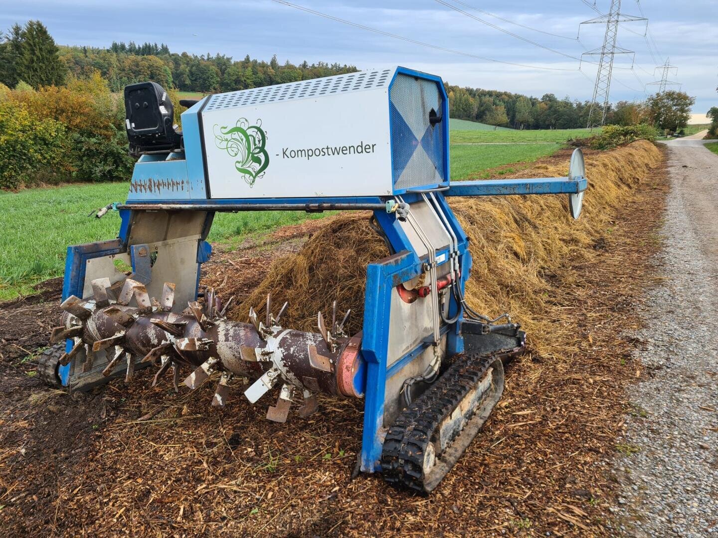 Ein blau-silberner Kompostwender steht auf einem Feldweg neben einem langen Kompost- und Mulchhaufen, im Hintergrund grüne Felder und Bäume unter einem bewölkten Himmel.