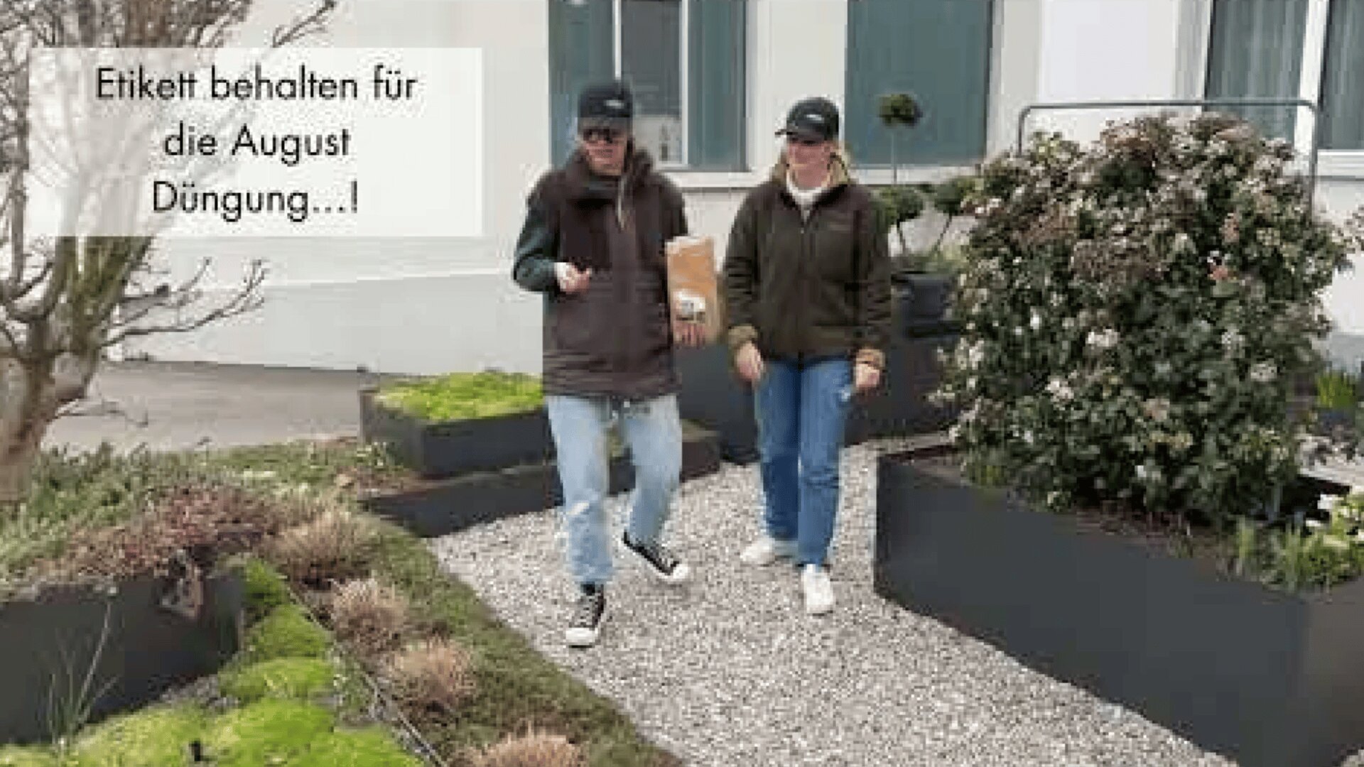 Zwei Personen mit legeren Jacken, Hüten und Sonnenbrillen gehen gemeinsam auf einem Kiesweg durch einen Garten mit Hochbeeten. Der deutsche Text auf dem Bild lautet: Etikett behalten für die August Düngung...!.