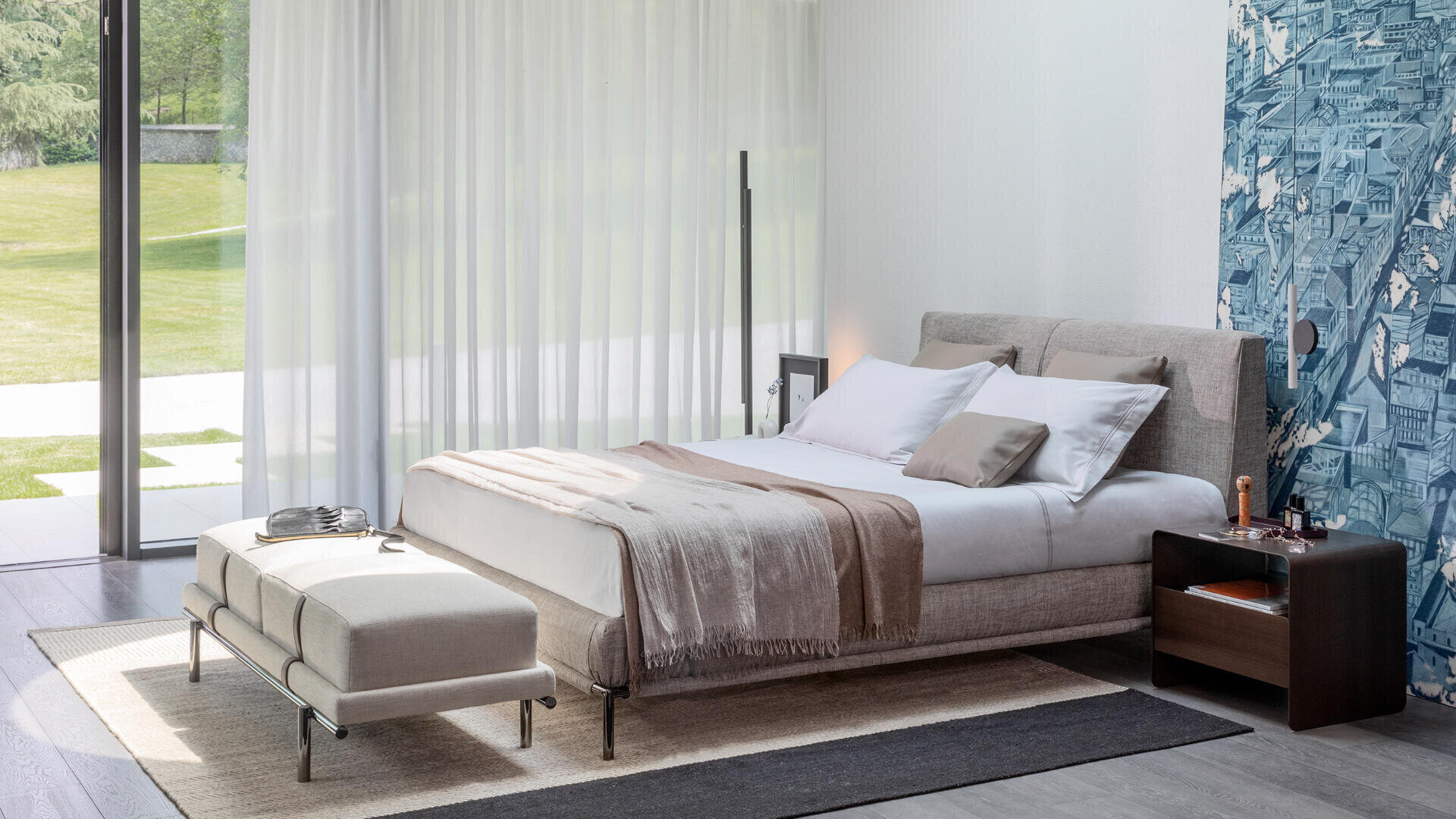 Ein modernes Schlafzimmer mit einem großen Bett, beiger Bettwäsche und einer neutralfarbenen Decke. Am Fußende des Bettes befindet sich eine Bank. Der Raum verfügt über raumhohe Fenster mit weißen Vorhängen und eine gemusterte blaue Akzentwand.