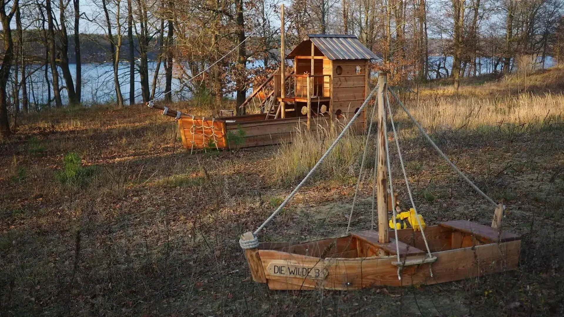 Two small wooden play boats and a playhouse sit in a grassy, wooded area near a lake. The boats and house are made from planks, and the area is surrounded by leafless trees on a sunny day.