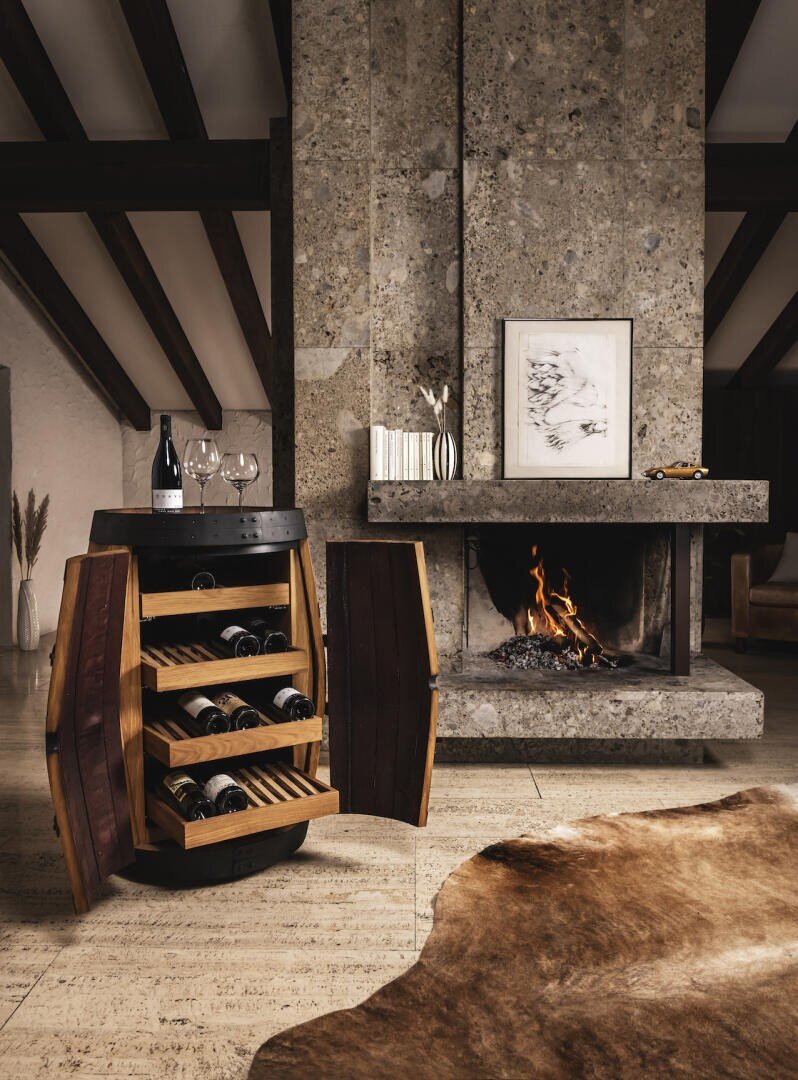 Ein Weinfassschrank mit offenen Türen stellt Weinflaschen und Gläser vor einem Steinkamin mit einem brennenden Feuer, gerahmten Kunstwerken darüber und einem Kuhfellteppich auf dem Boden in einem gemütlichen Wohnzimmer aus.