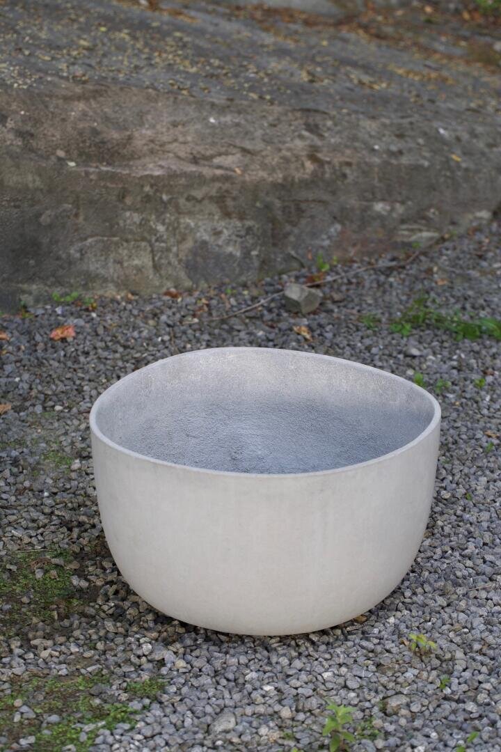 A large, round, white ceramic or concrete bowl sits outdoors on gravel, with a rocky wall and scattered greenery in the background.