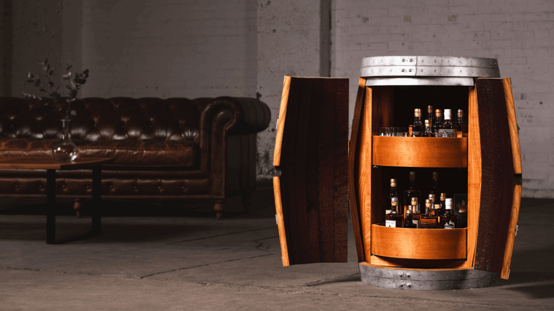 A wooden barrel cabinet with open doors displays shelves of liquor bottles inside. In the background, there is a brown leather sofa and a table with a vase, set in an industrial-style room with concrete floors and white brick walls.
