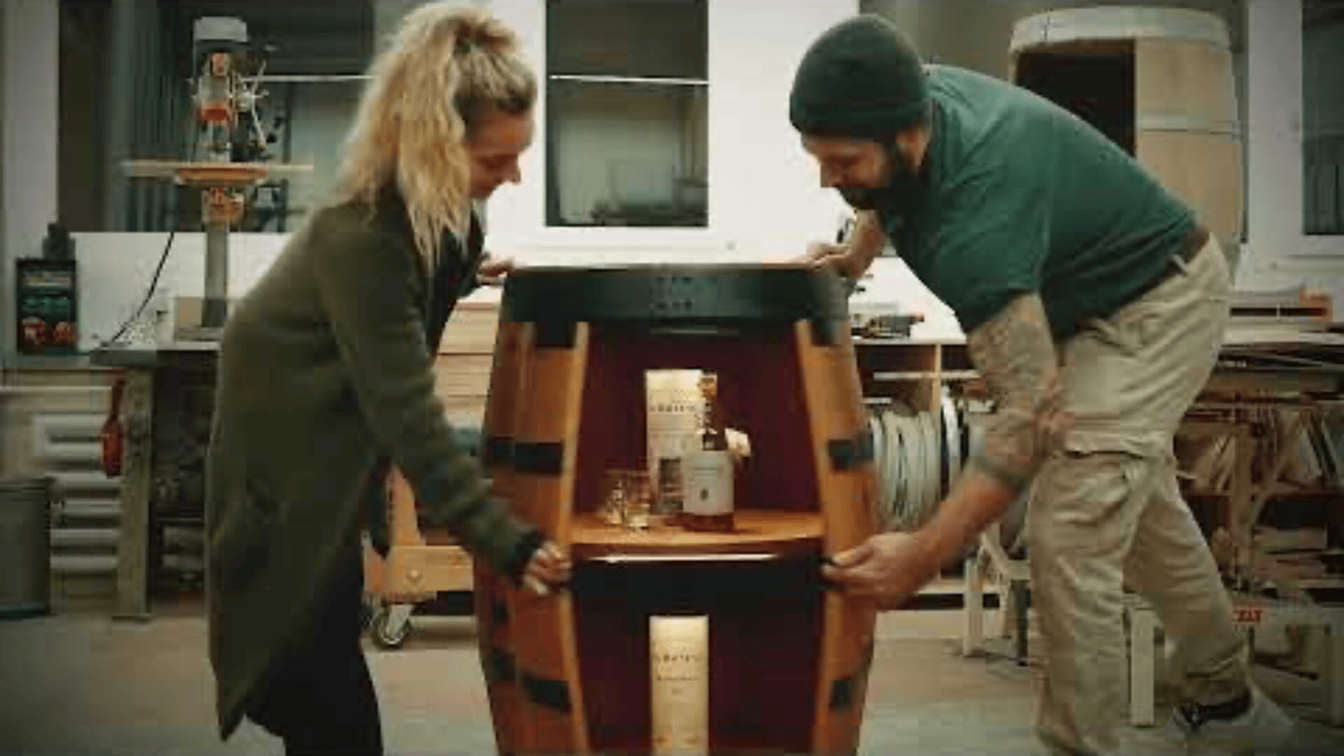 A woman and a man in a workshop are smiling as they move a wooden barrel that has been converted into a cabinet, revealing bottles and glasses stored inside.