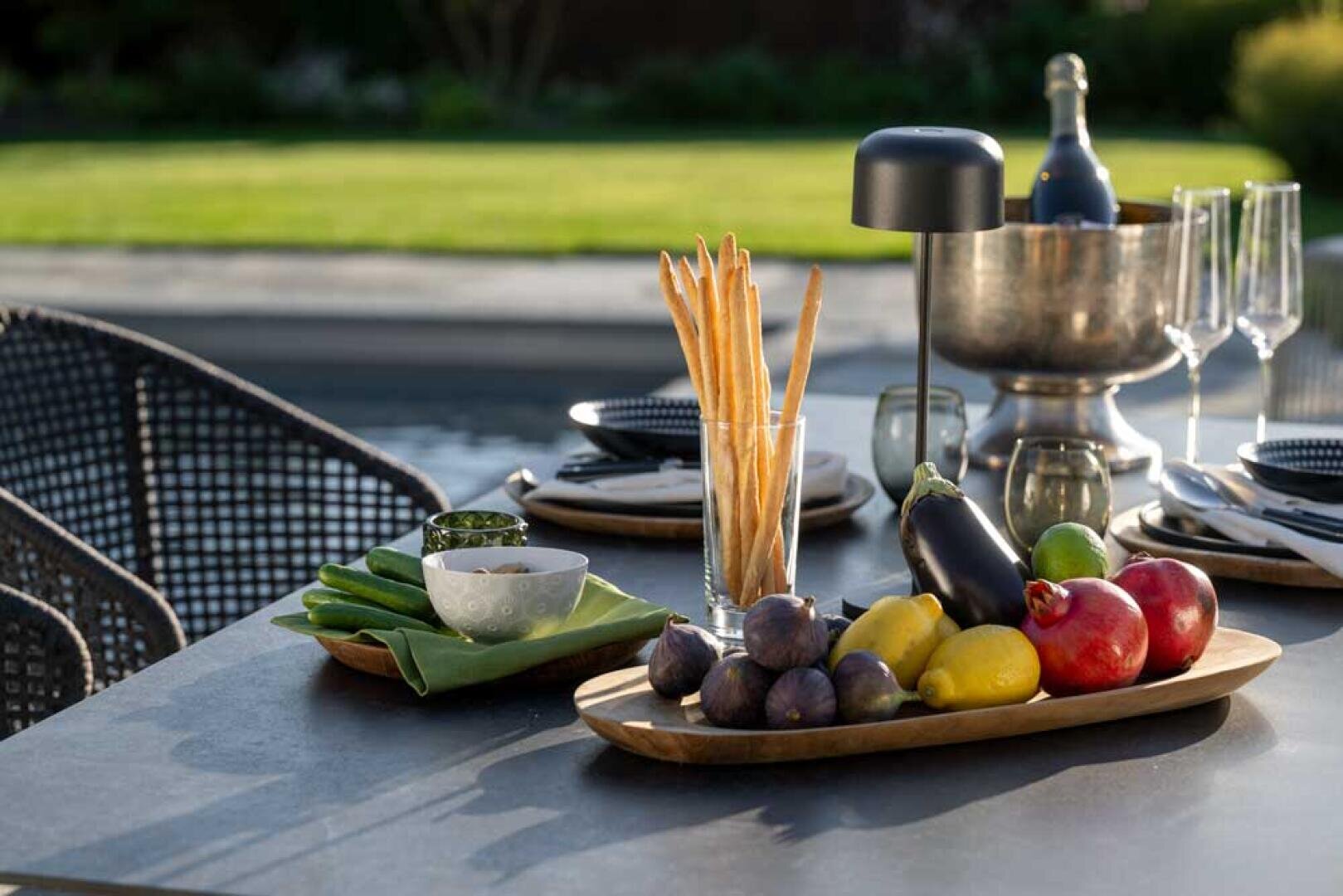 Ein moderner Esstisch im Freien mit Tellern, Gläsern, Brotstangen in einem Glas, verschiedenen frischen Früchten und Gemüse auf einem Holztablett und einer Weinflasche in einem Eiskübel aus Metall, mit einem Garten im Hintergrund.