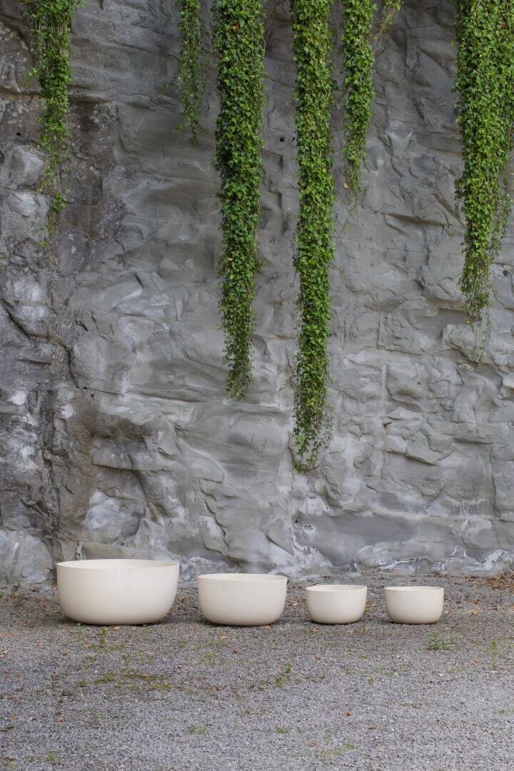 Four cream-colored round pots of different sizes are lined up on a gravel surface in front of a gray stone wall, with long green vines hanging down from above.
