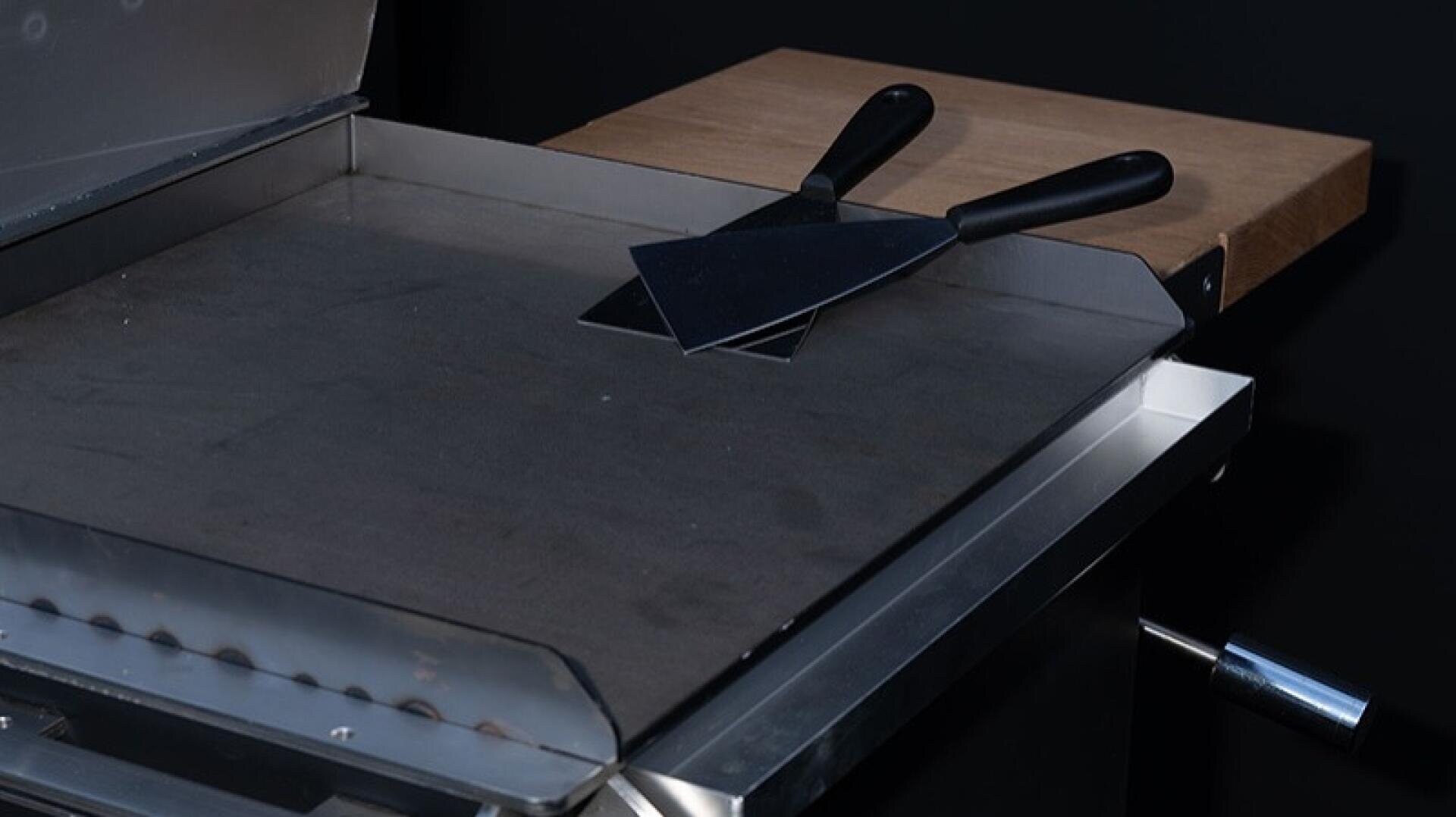 A metal griddle with two flat spatulas crossed on top, featuring black handles. The griddle has a wooden side shelf and is set against a dark background.