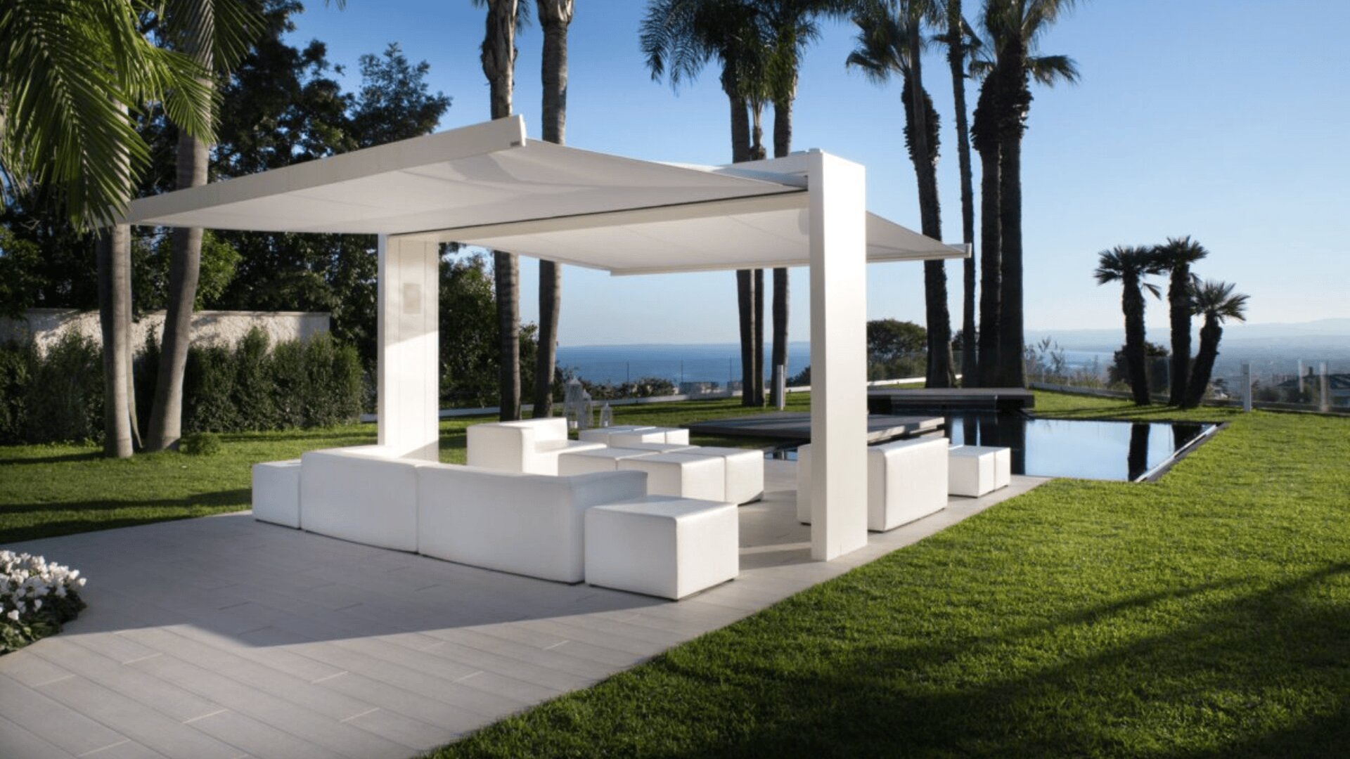 Auf der Terrasse neben dem rechteckigen Pool, umgeben von Gras und Palmen und mit Blick auf das Meer im Hintergrund, befindet sich ein moderner Lounge-Bereich mit weißen Sitzmöbeln und einem Baldachin.