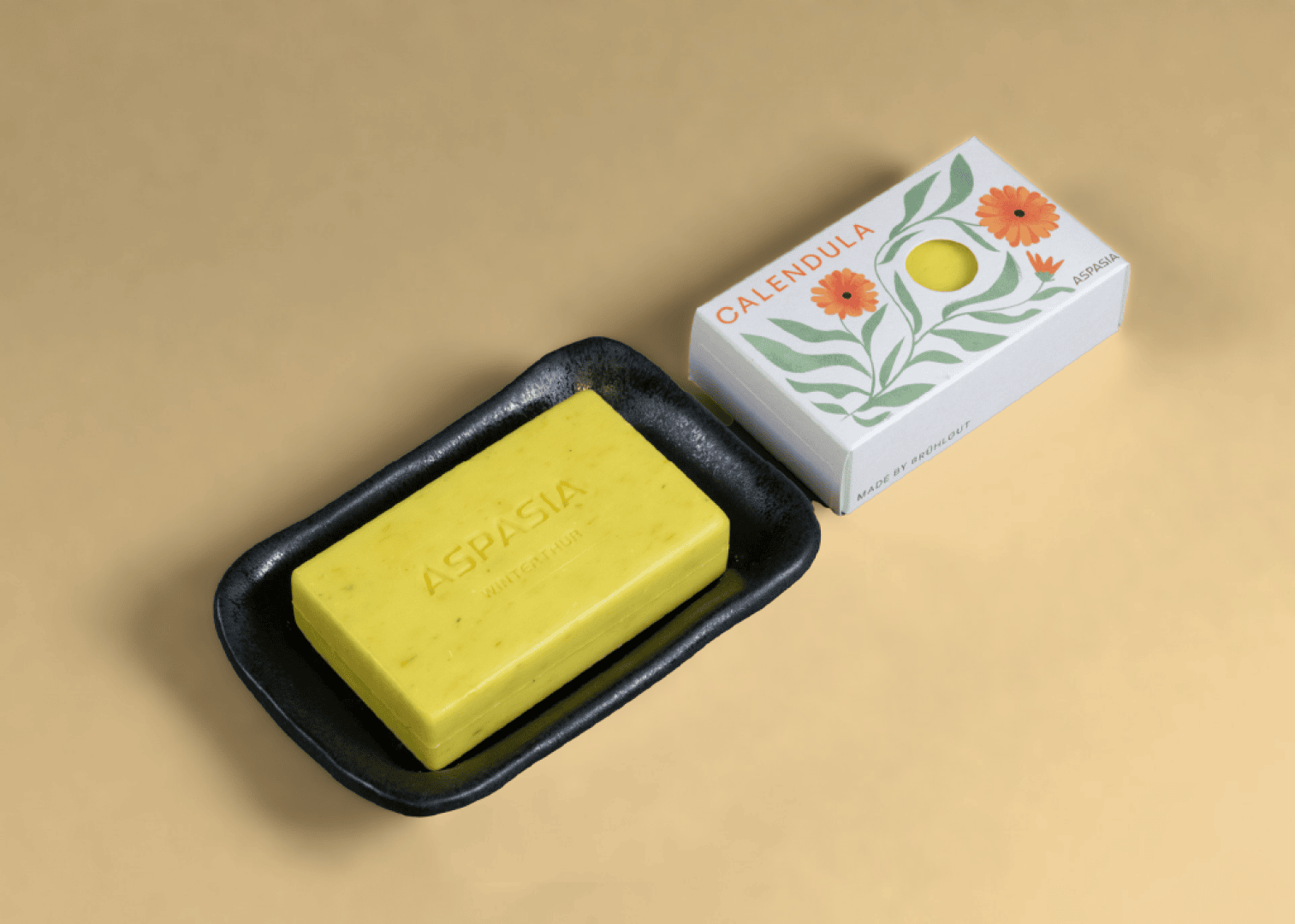 Ein gelbes Seifenstück mit der Aufschrift ASPASIA liegt in einer schwarzen Seifenschale neben einer weißen Schachtel mit orangefarbenen Blüten und grünen Blättern, die die Aufschrift CALENDULA trägt.